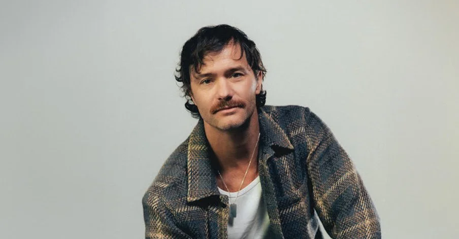 A man with dark wavy hair, a mustache, and light skin, wearing a plaid jacket over a white shirt, looking at the camera against a plain background.