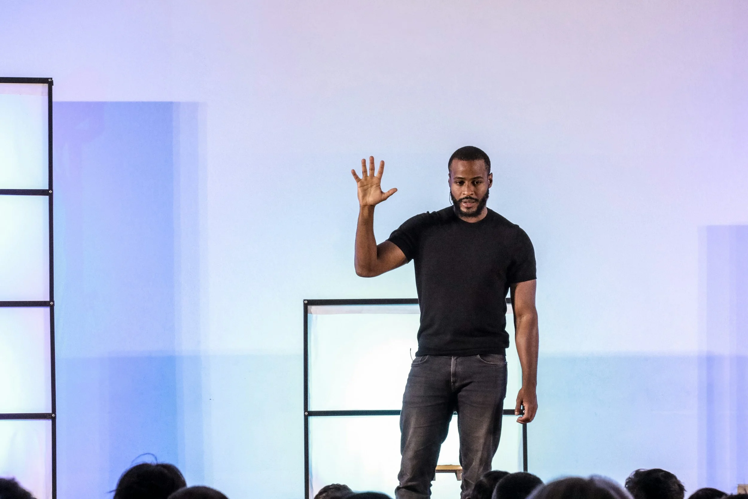 A man in a black t-shirt and gray jeans standing on stage with a colorful background, raising his right hand as he speaks to an audience.
