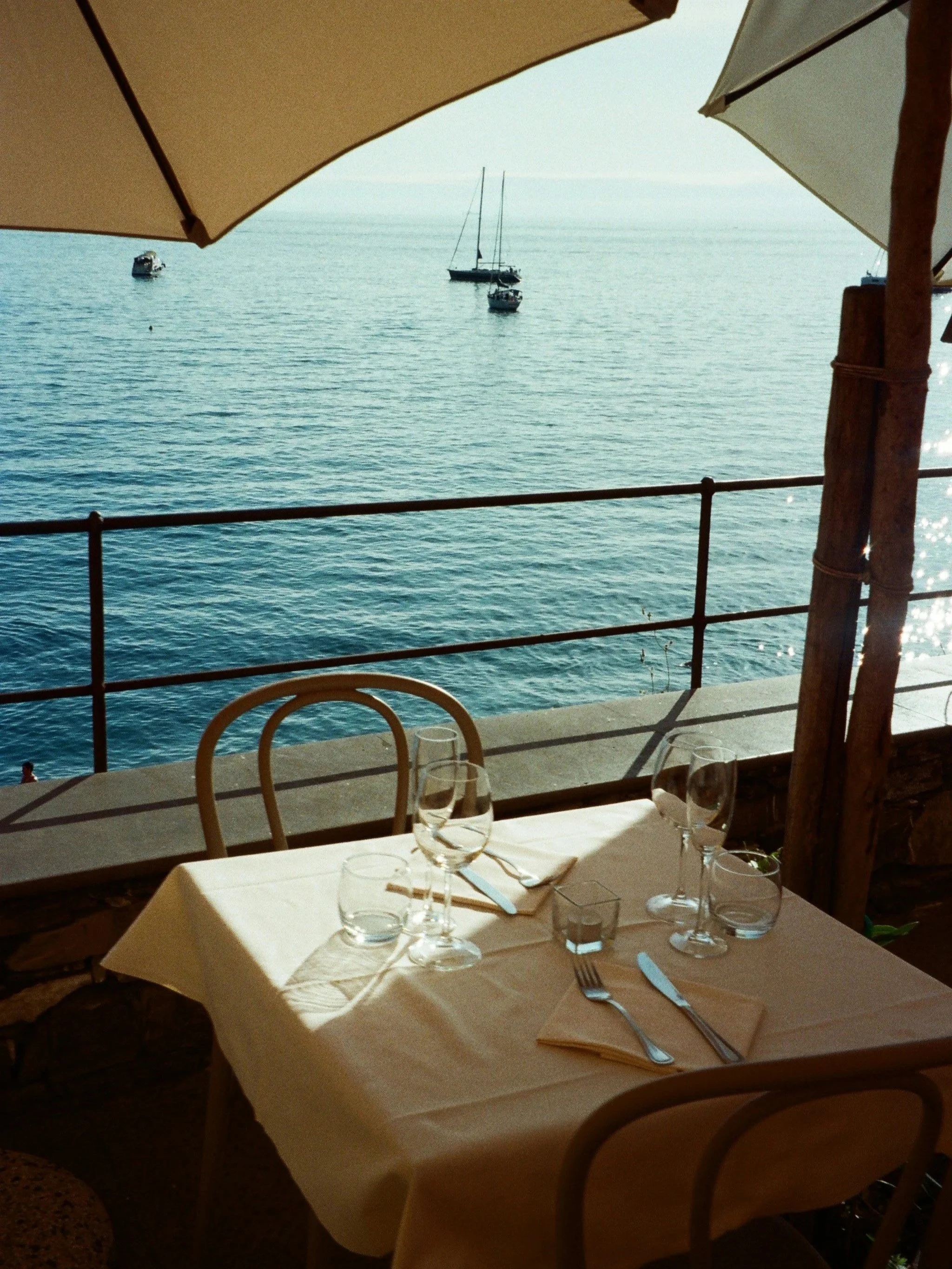 A dining table set for two with white napkins, wine glasses, and water glasses, overlooking a calm sea with sailboats.