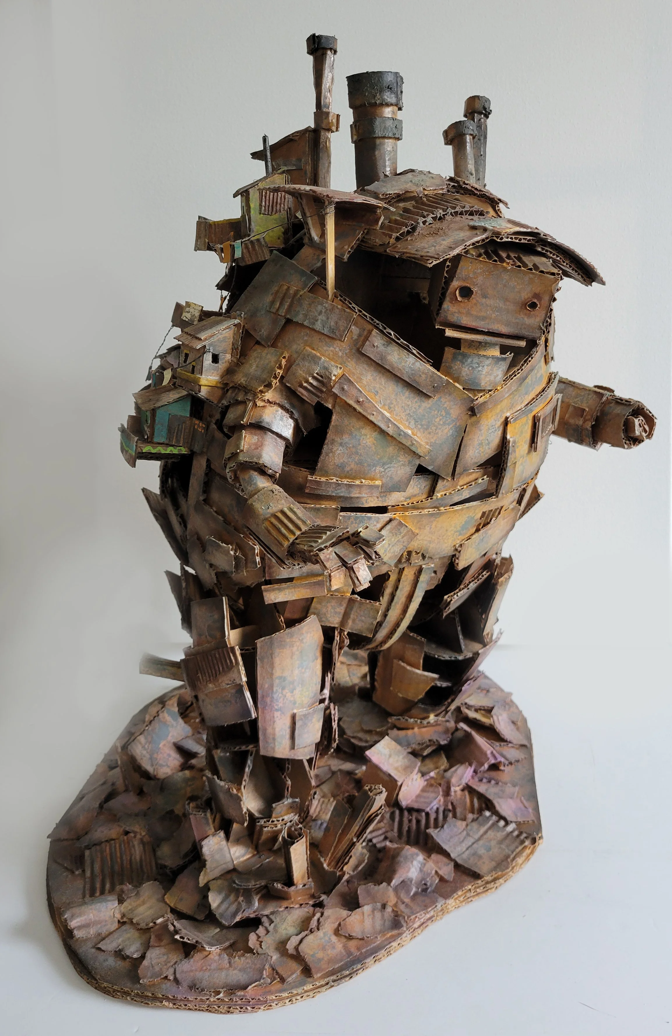 Sculpture of a robot or humanoid figure made from layered, rusted corrugated cardboard and metal pieces, with a rounded base and four smokestacks on its head, against a plain white background.