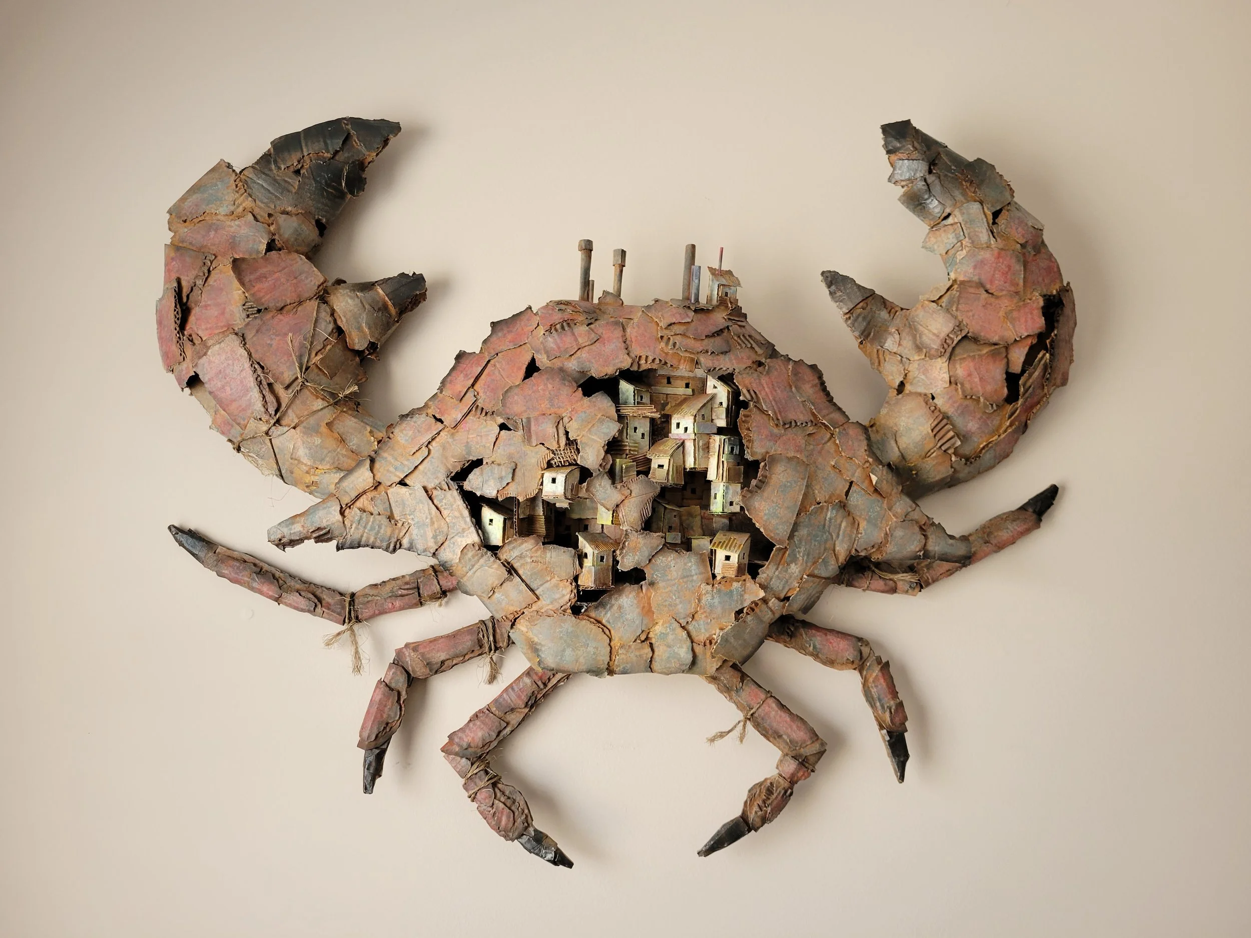 A wall art piece depicting a crab made from cardboard, with the body consisting of small building models.