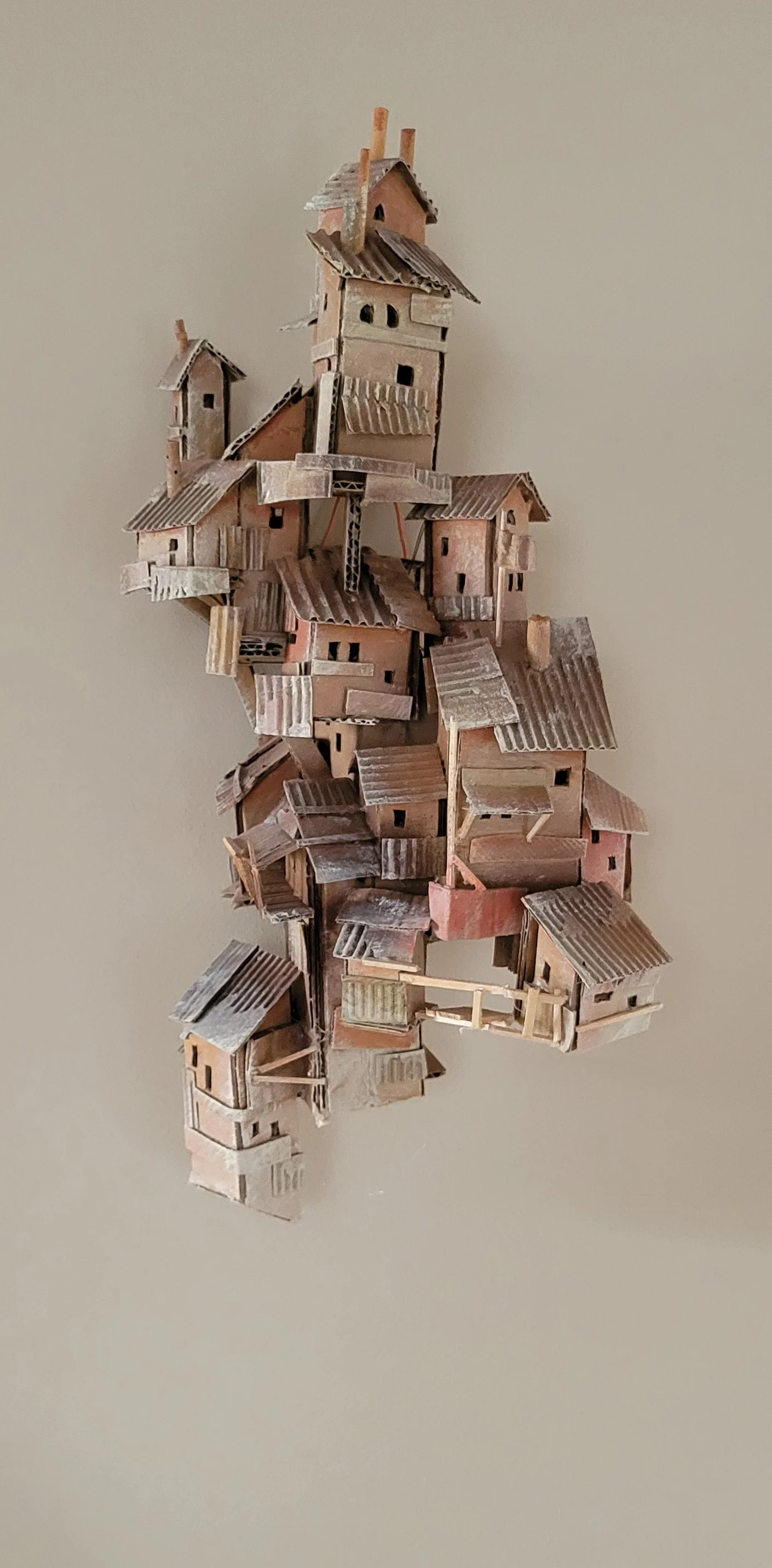 A vertical miniature model of a multi-story house complex made from cardboard and wood, with detailed roofs, windows, and balconies, set against a plain background.