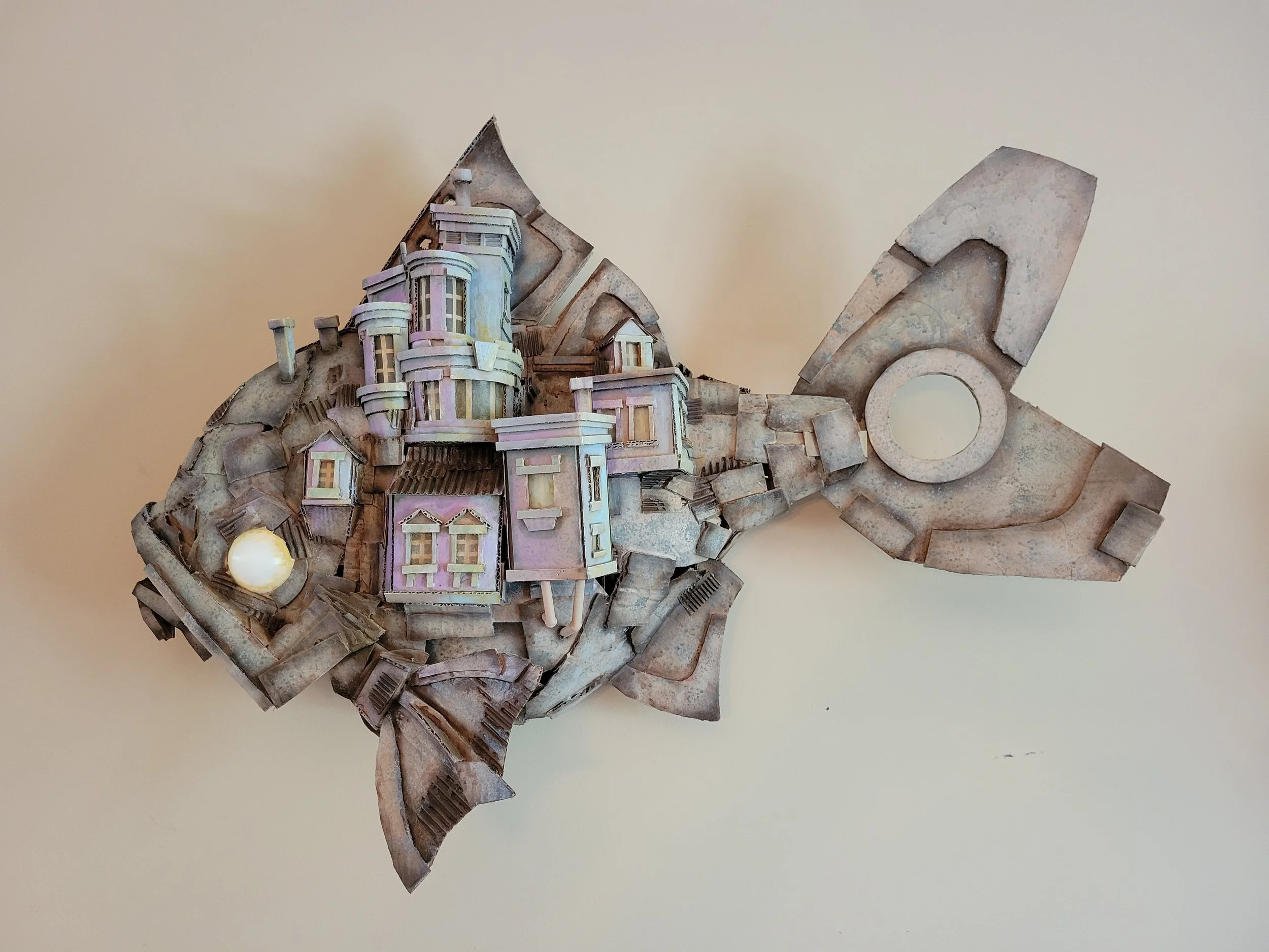 A sculpture of a fish made of cardboard, with a small house structure attached.