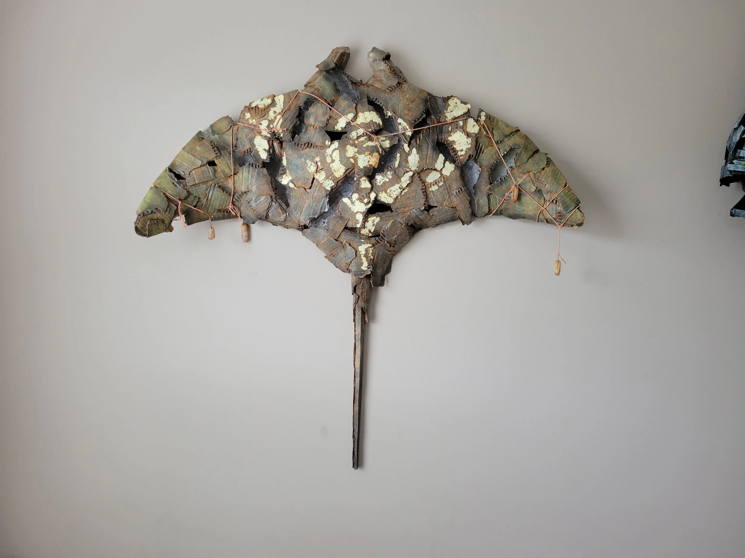 A wall decoration made of cardboard assembled in the shape of a sting ray, decorated with small hanging shells and wrapped with thin wires.