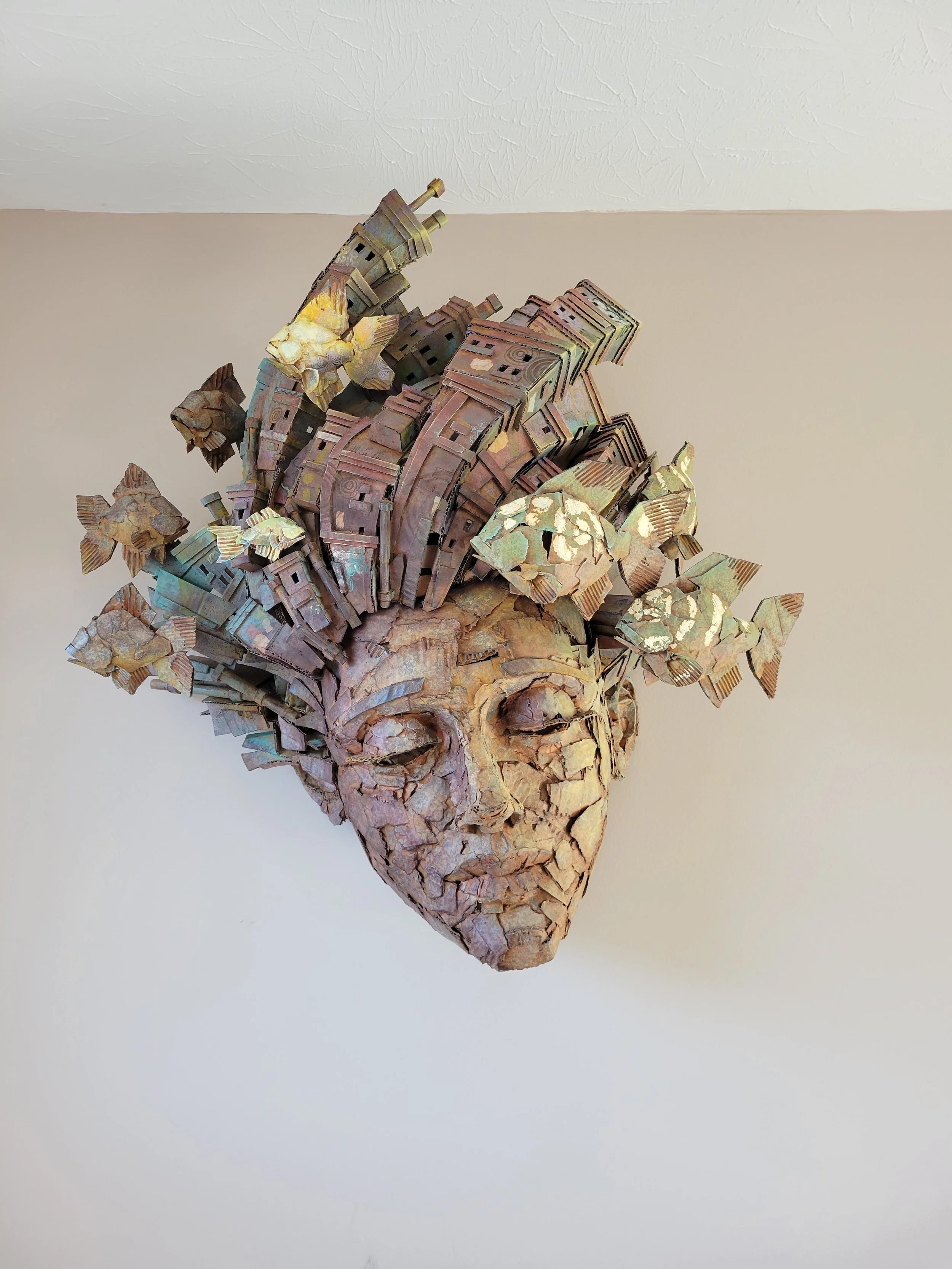 Mixed media sculpture of a woman's face with city-like structures emerging from the top, set against a plain background.