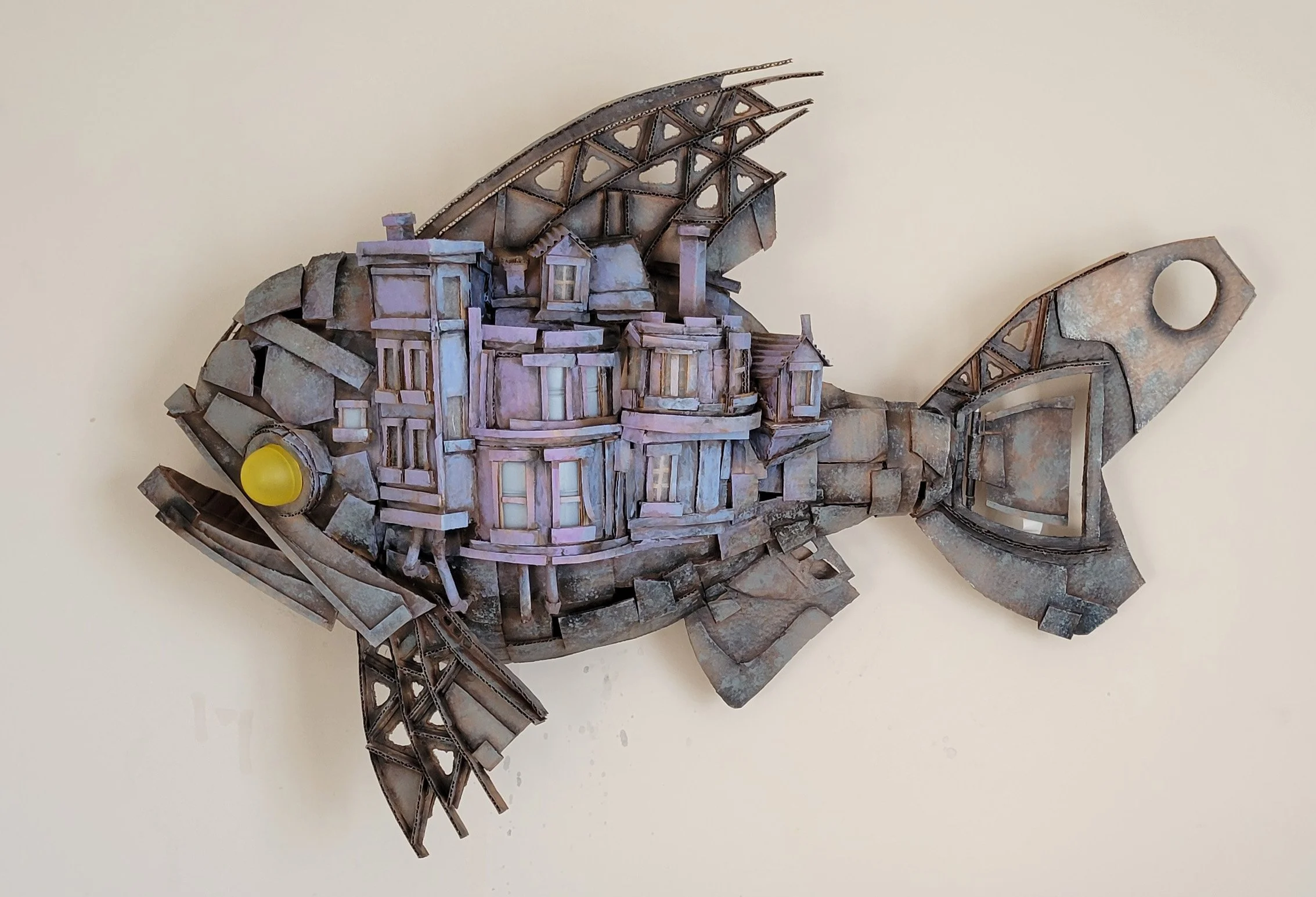 A metal sculpture of a fish made from various scrap metal parts, with detailed scales, fins, and a large eye, mounted on a plain wall.