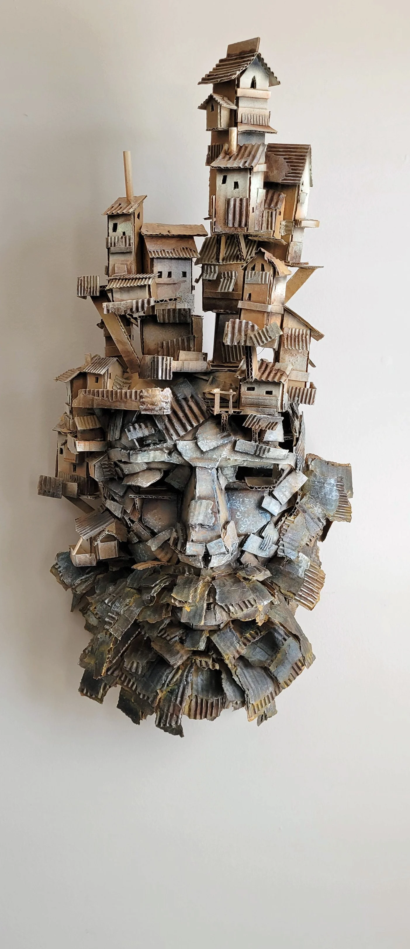 Mixed media sculpture of an elderly man's face with a crown of buildings and rooftops on his head, made from various small architectural elements and materials.