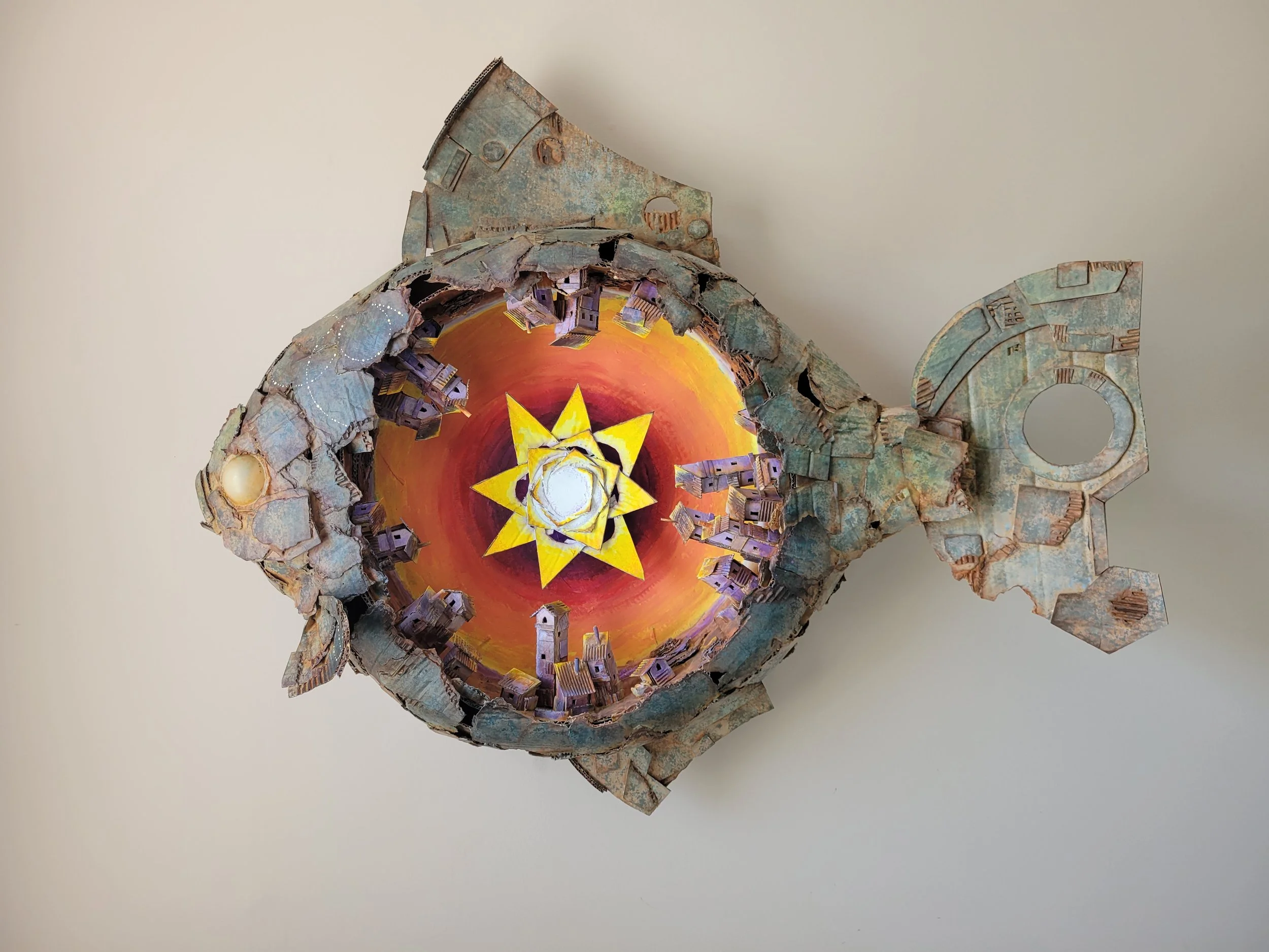 A mixed media art piece resembling a fish with an inner painted scene of a colorful swirling sunset with a yellow star in the center, surrounded by small model houses and buildings, crafted from cardboard.