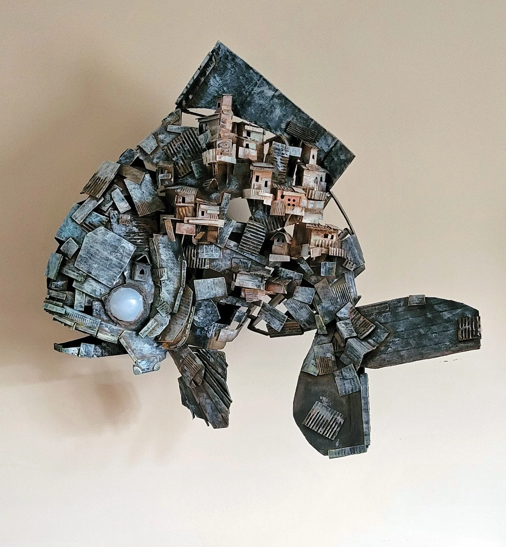 A sculpture of a fish constructed from various cardboard pieces, including small buildings and other recycled materials.