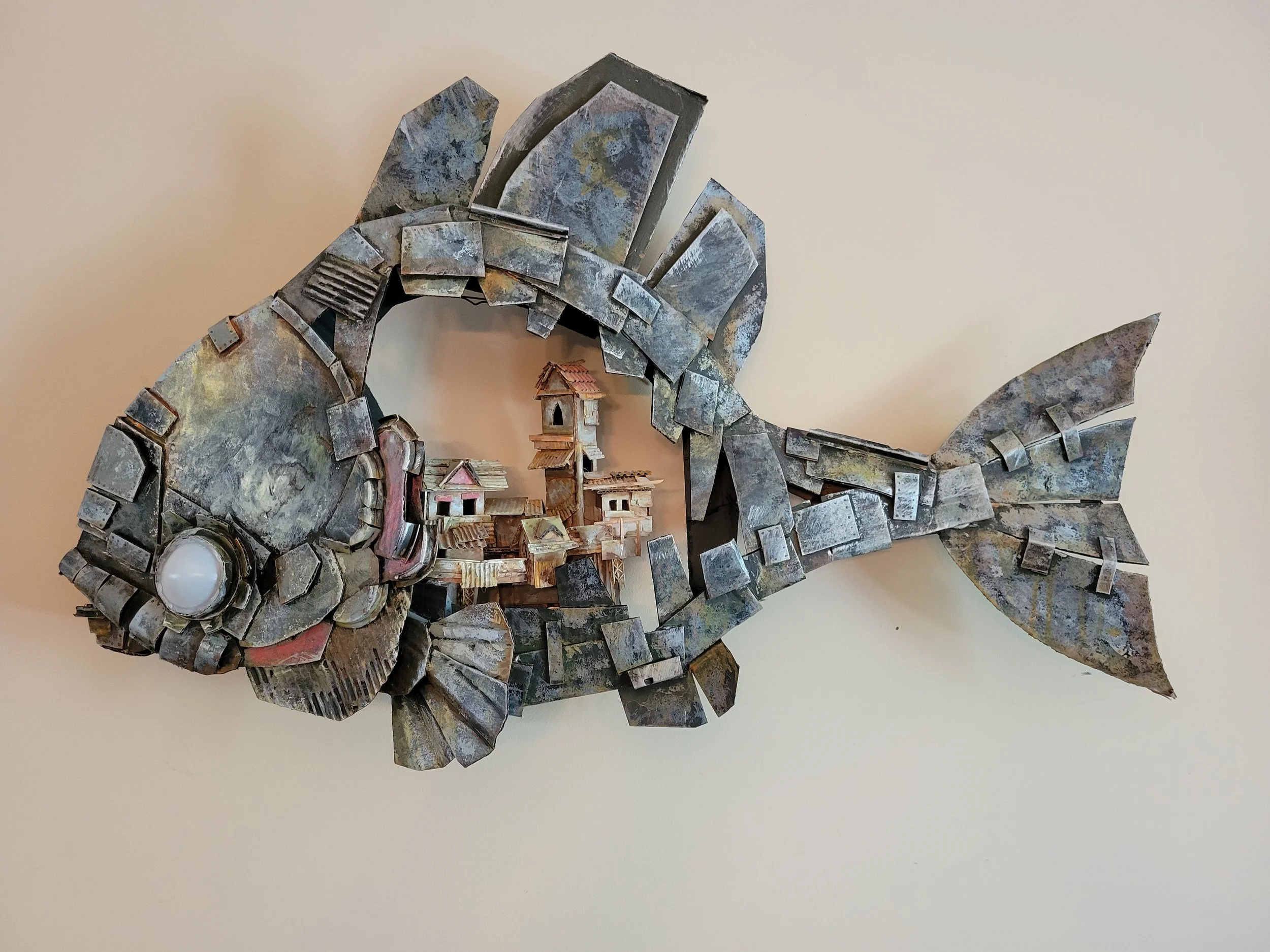 A wall sculpture of a fish made from cardboard, with miniature houses built in.