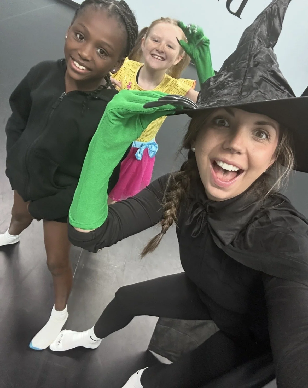 Three young girls dressed in costumes, with one wearing a witch hat and a black cape, taking a selfie together.