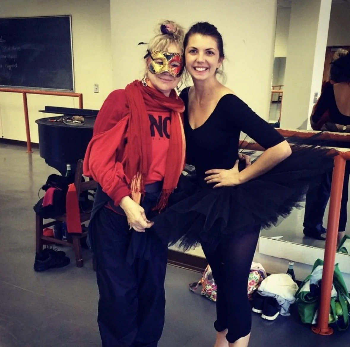 Two women in a room, one dressed in a red jacket, red scarf, black pants, wearing a colorful mask and the other in a black dance outfit with a tutu, smiling together. The woman on the right has her hand on her hip. In the background, a blackboard, a black piano, and some bags and shoes on the floor are visible.