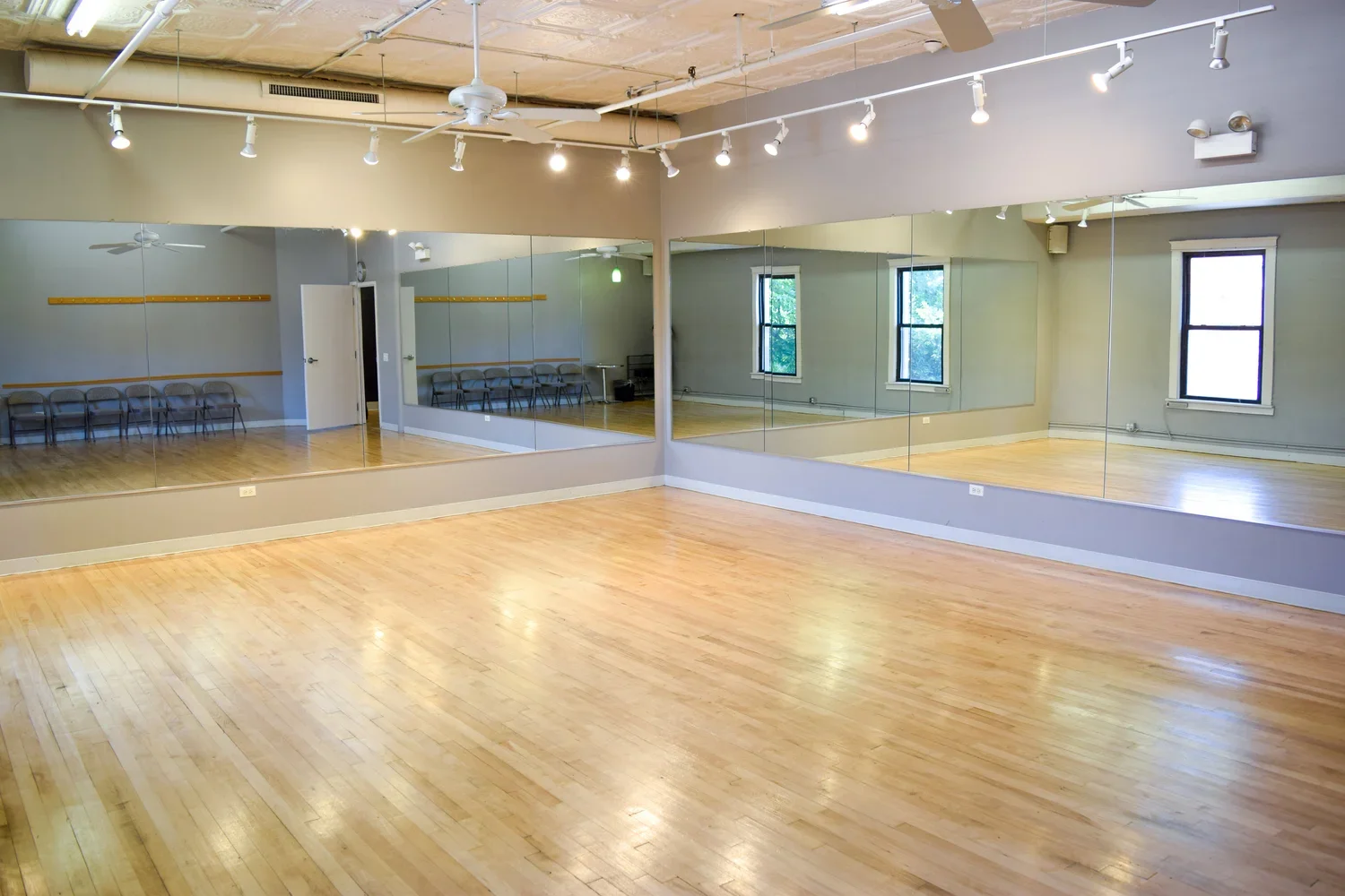 Empty dance studio with wooden floors, a large mirror wall, windows, and chairs along the wall.