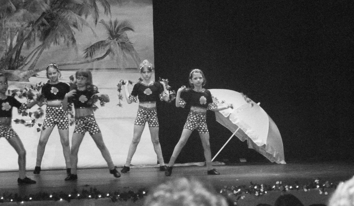 Group of young girls performing a dance on stage, dressed in coordinated outfits with floral tops and checkered shorts, holding flowers and an umbrella, with a tropical beach backdrop painted on the wall behind them.