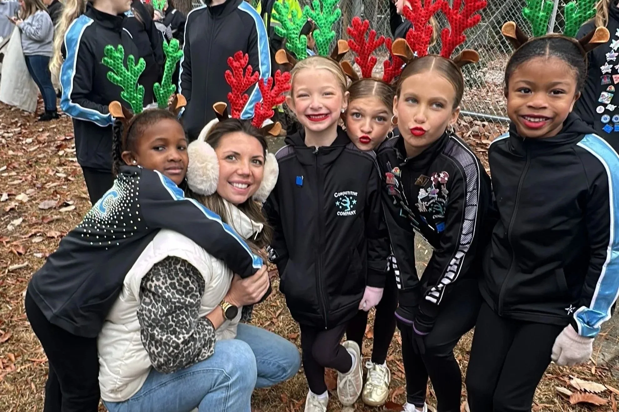A group of children and an adult woman wearing Christmas-themed reindeer antlers headbands, posing outdoors on a leaf-covered ground, with some children making funny faces and others smiling.