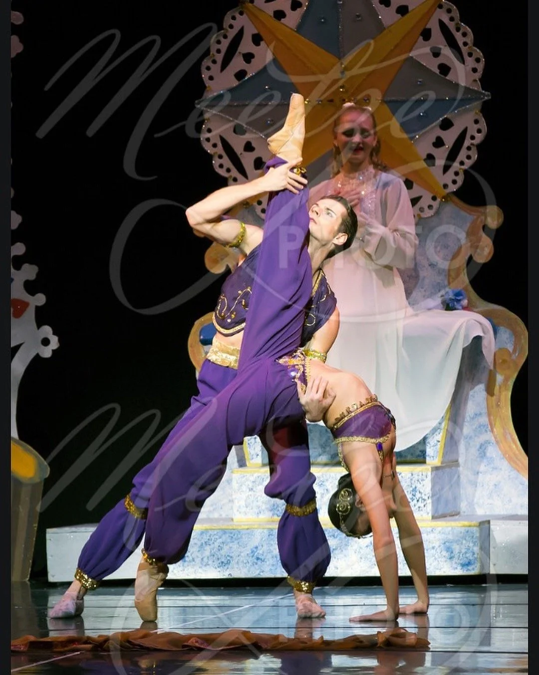 Two ballet dancers performing in costume on stage, with one bending backward and the other supporting, and a woman seated in the background watching.
