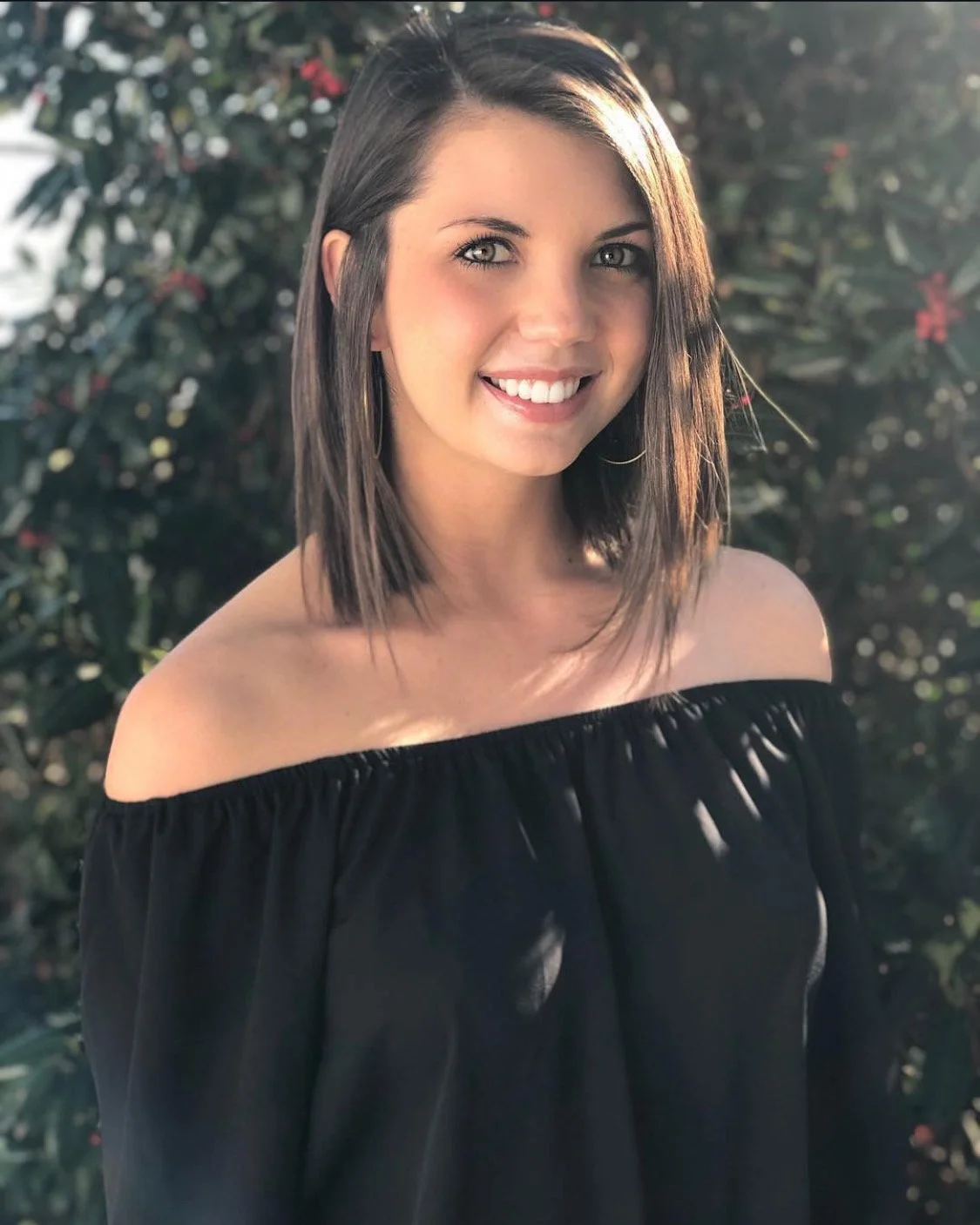 Young woman with shoulder-length brown hair, smiling in front of greenery, wearing an off-the-shoulder black top.