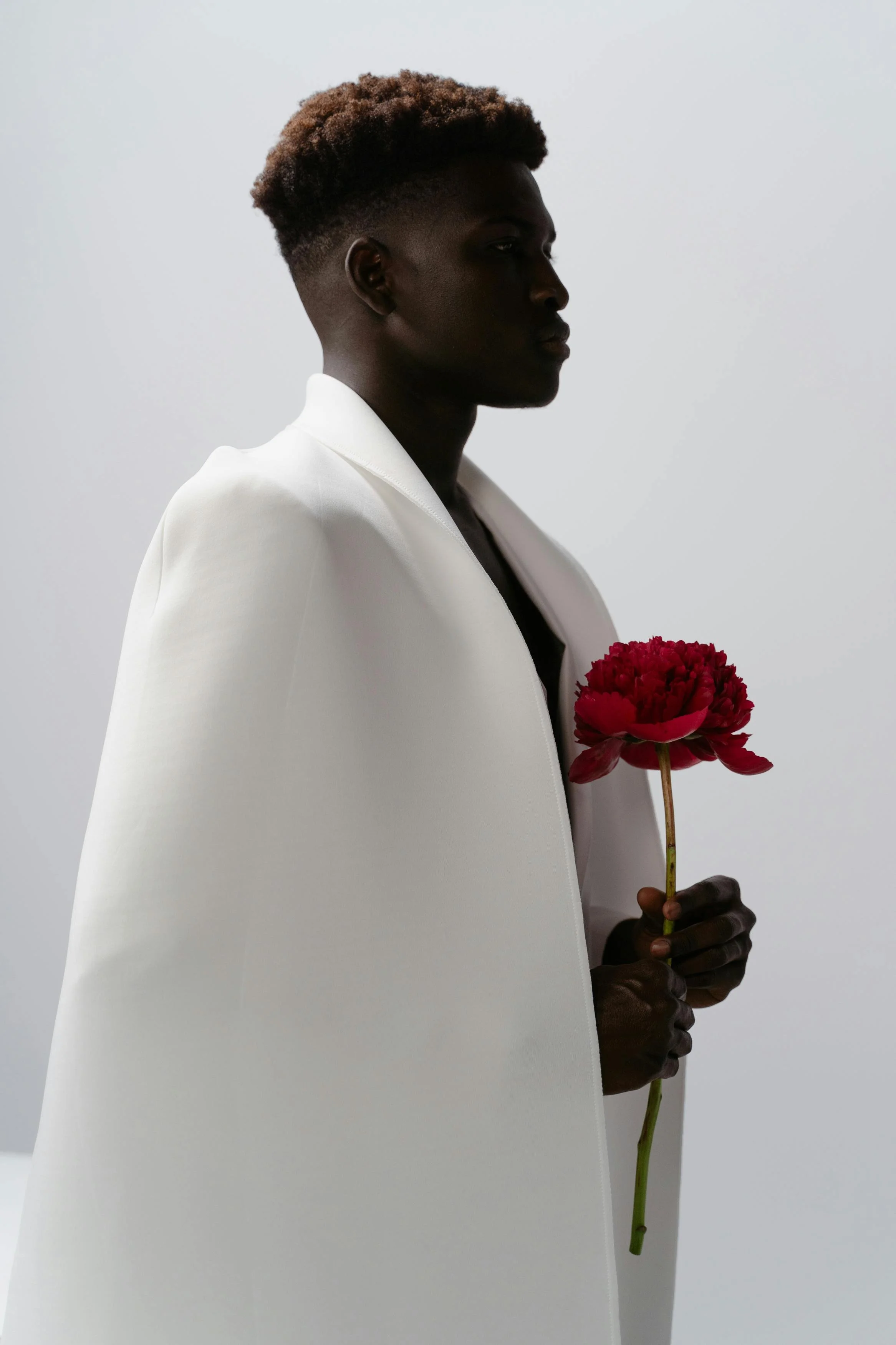 Person with dark skin wearing a white coat, holding a red flower, standing against a plain light background.