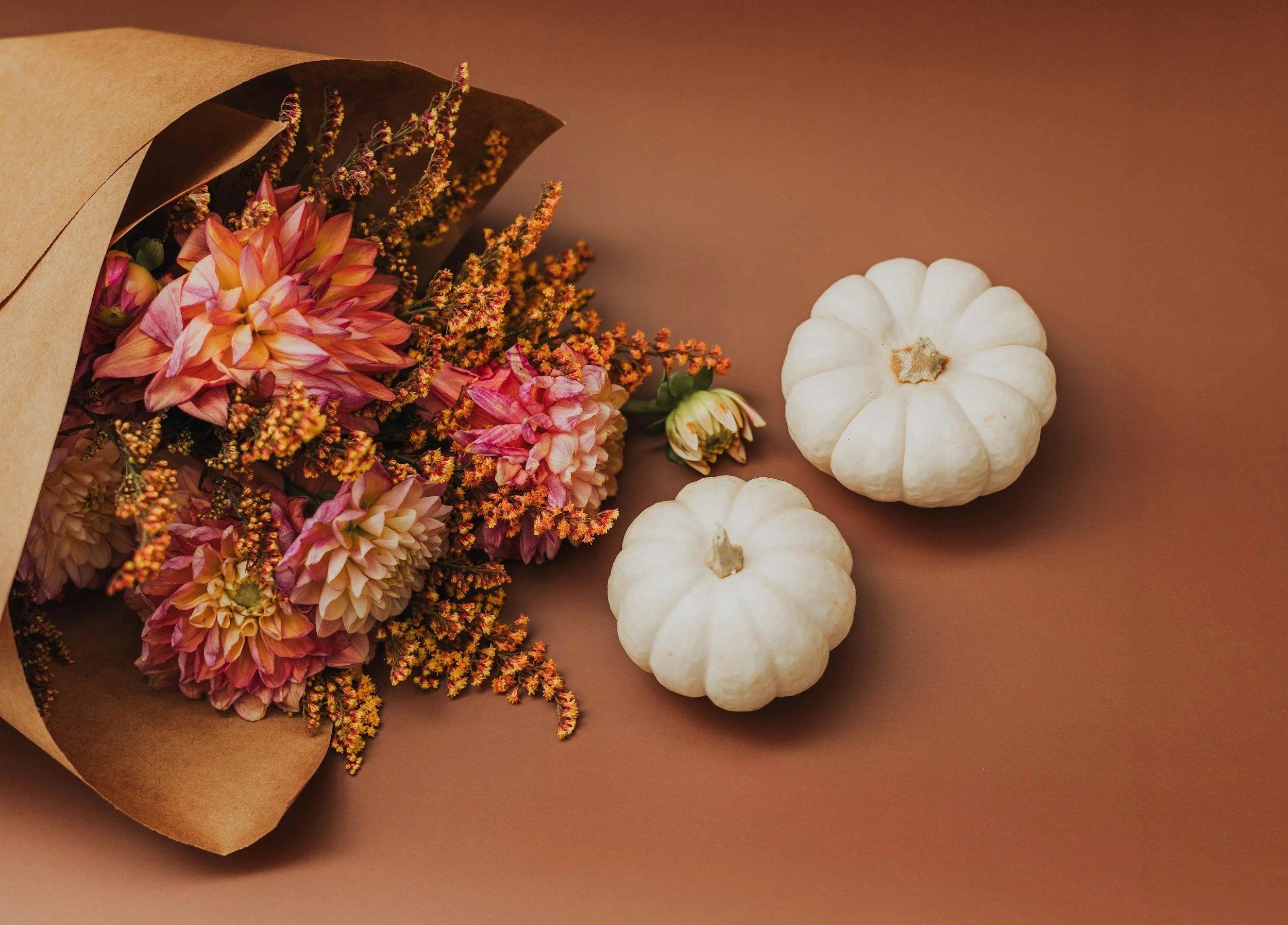 Bouquet of pink and orange dahlias and yellow flowers wrapped in brown paper, accompanied by two small white pumpkins on a brown background.