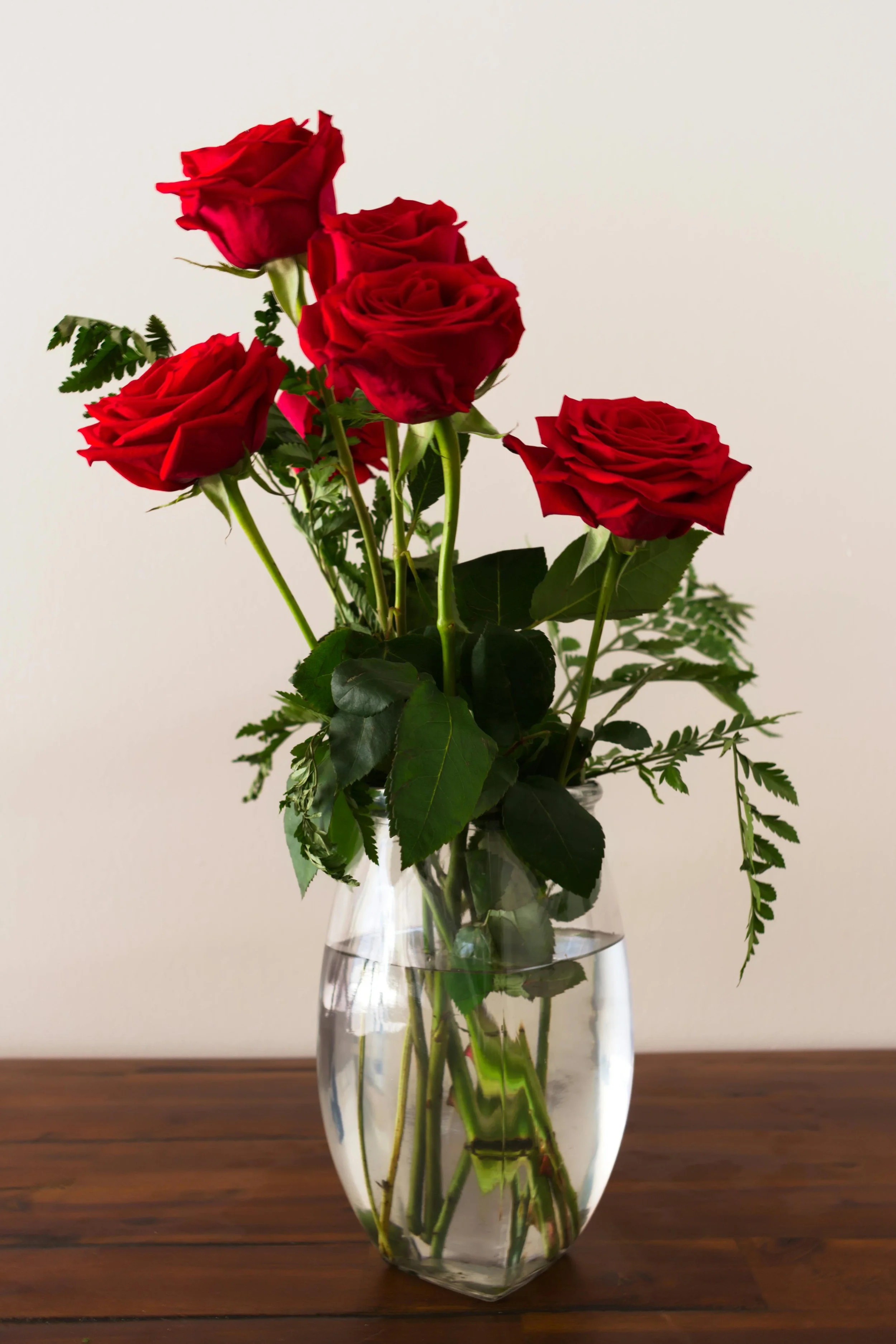 A bouquet of red roses and green foliage in a clear glass vase on a wooden surface with a plain white background.