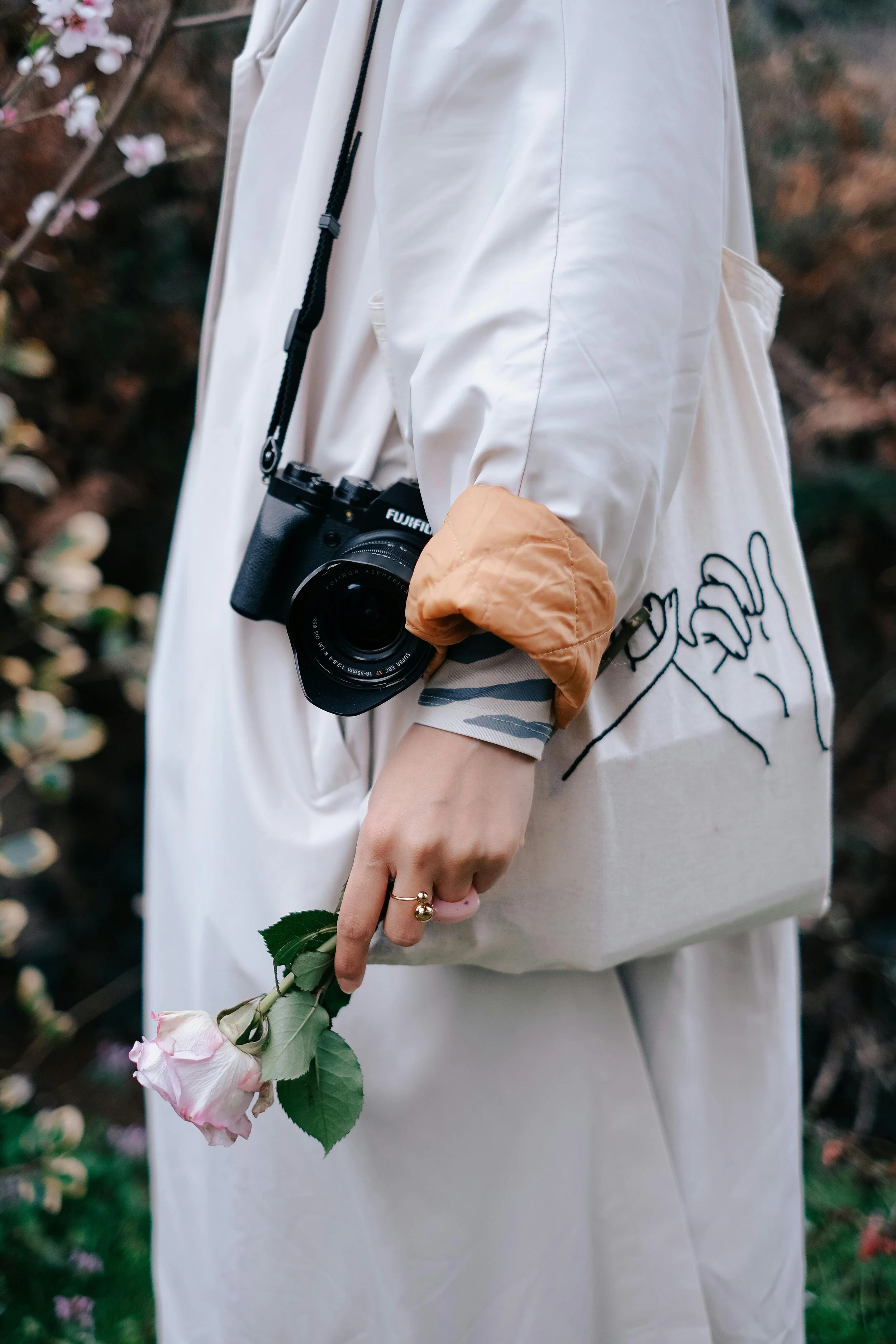 Person wearing a white coat with a beige and gray sleeve, holding a pink rose with green leaves, and carrying a beige tote bag with black drawing. A black camera hangs around their neck. The background shows blurred plants with pink flowers.