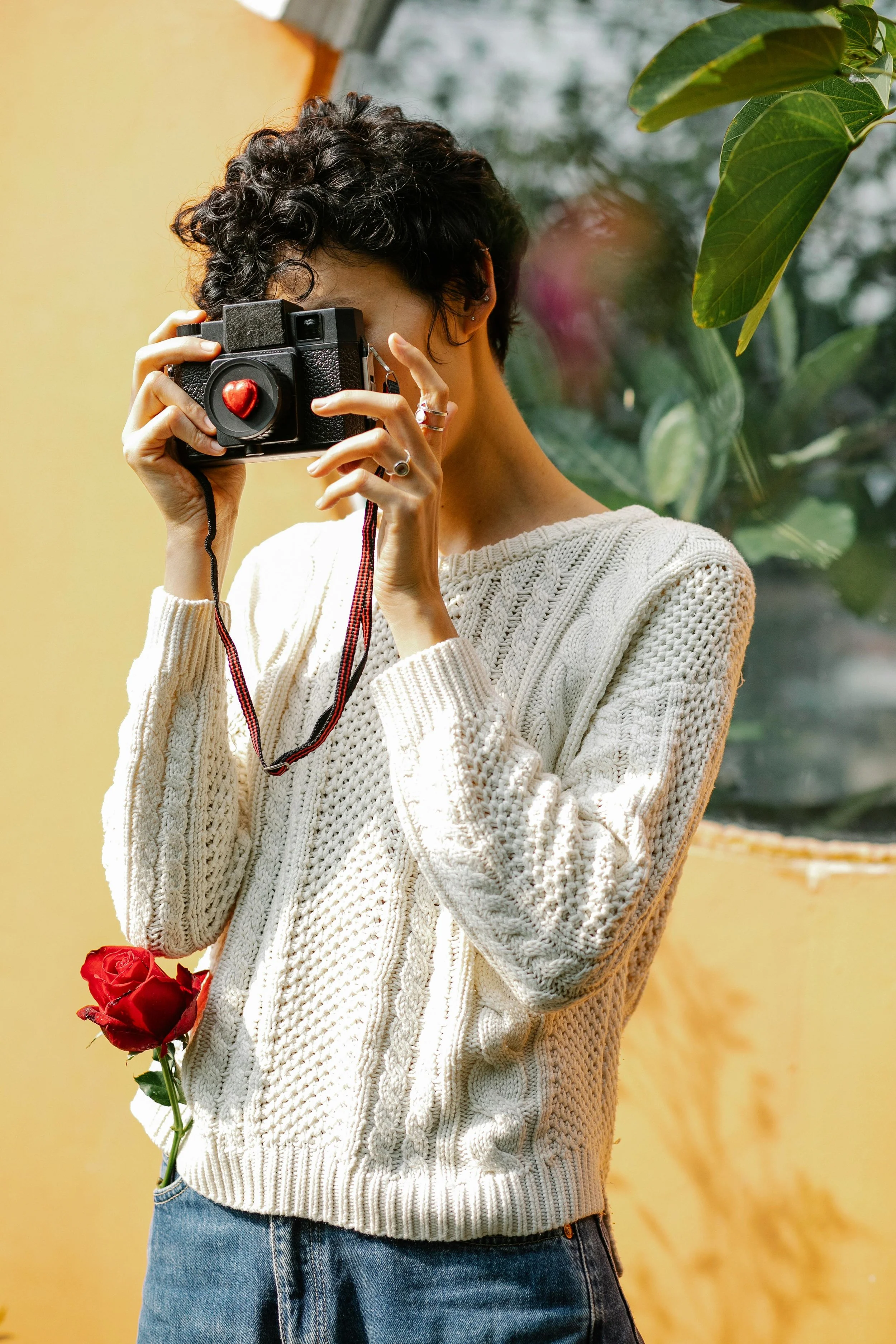 A woman with short, curly hair takes a photo with a camera that has a red heart-shaped decoration on the front, while a red rose is tucked into her pocket.