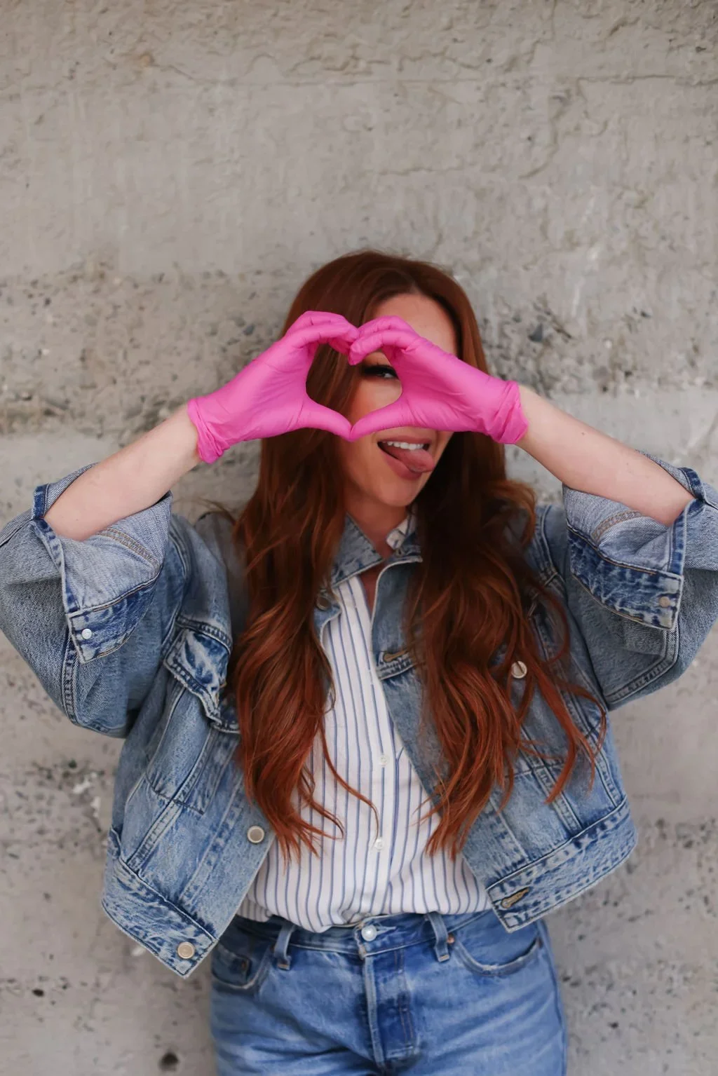 A woman with long red hair, wearing a denim jacket and jeans, pasting pink gloves, making a heart shape with her hands, and sticking out her tongue.