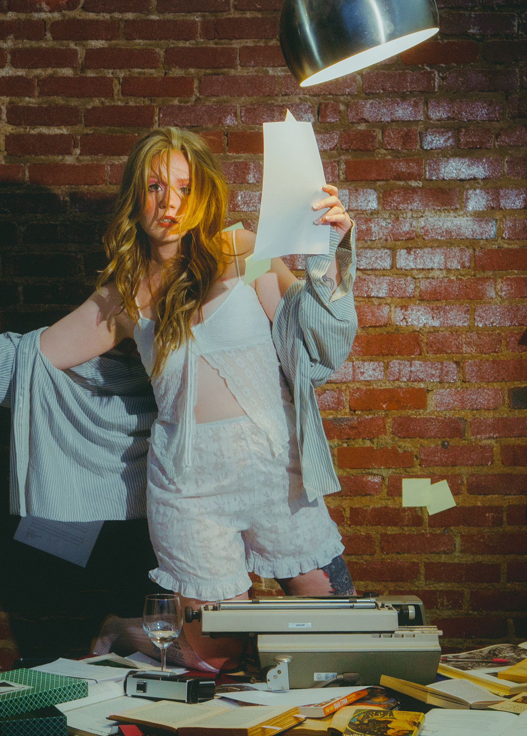 A woman with long, wavy hair and a tattoo on her left thigh, standing against a brick wall, holds a piece of paper near her face with a typewriter in front of her. The table is cluttered with books, papers, a glass of wine, and various crafting supplies, with a metallic lamp casting light from above.