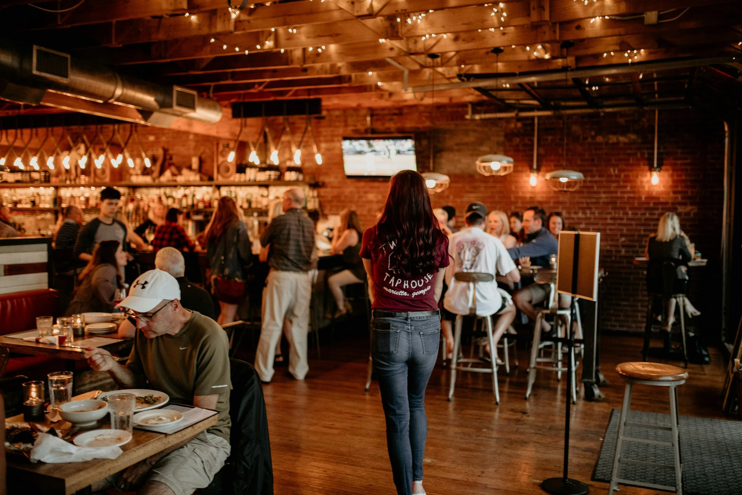 A woman with long dark hair standing in a rustic restaurant or bar interior with exposed brick walls and dim lighting, overlooking groups of people dining and socializing.