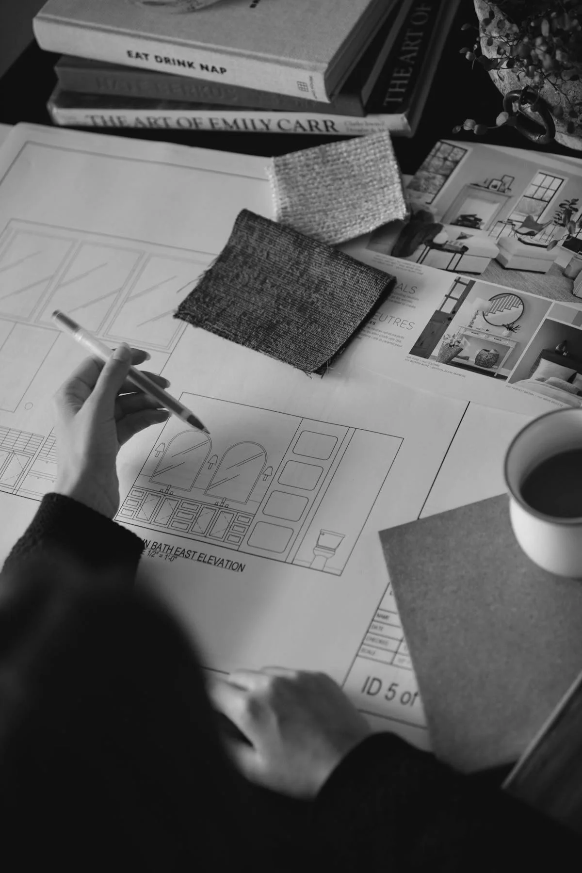 A person working on interior design plans at a desk with fabric samples, design magazines, and architectural drawings, along with a cup of coffee.