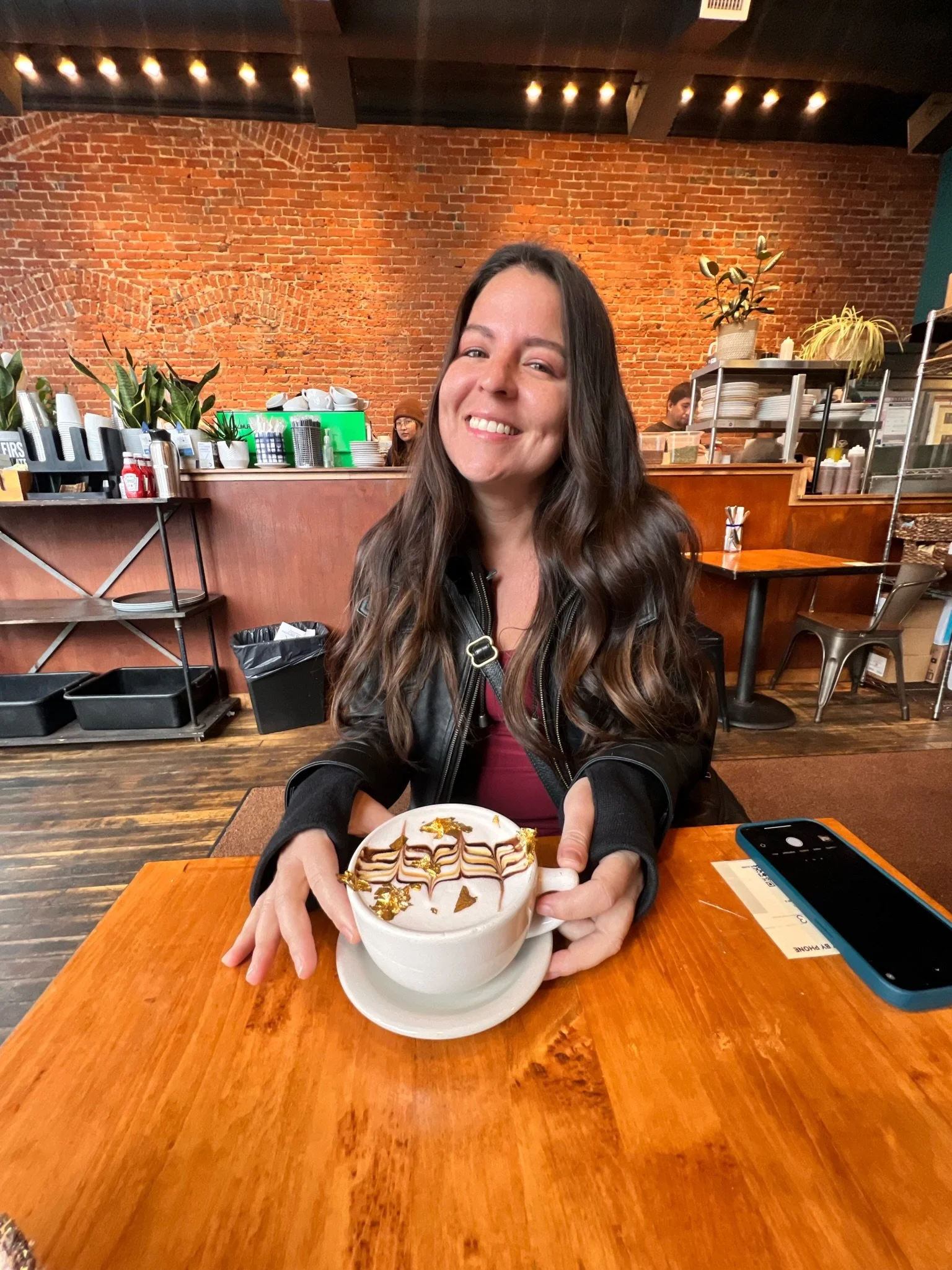 A smiling woman with long brown hair wearing a black leather jacket is sitting in a café. She is holding a large cappuccino with whipped cream, chocolate drizzle, and gold leaf decoration. The café has a brick wall background, with plants, plates, and cups on shelves, and other customers and staff visible in the background.
