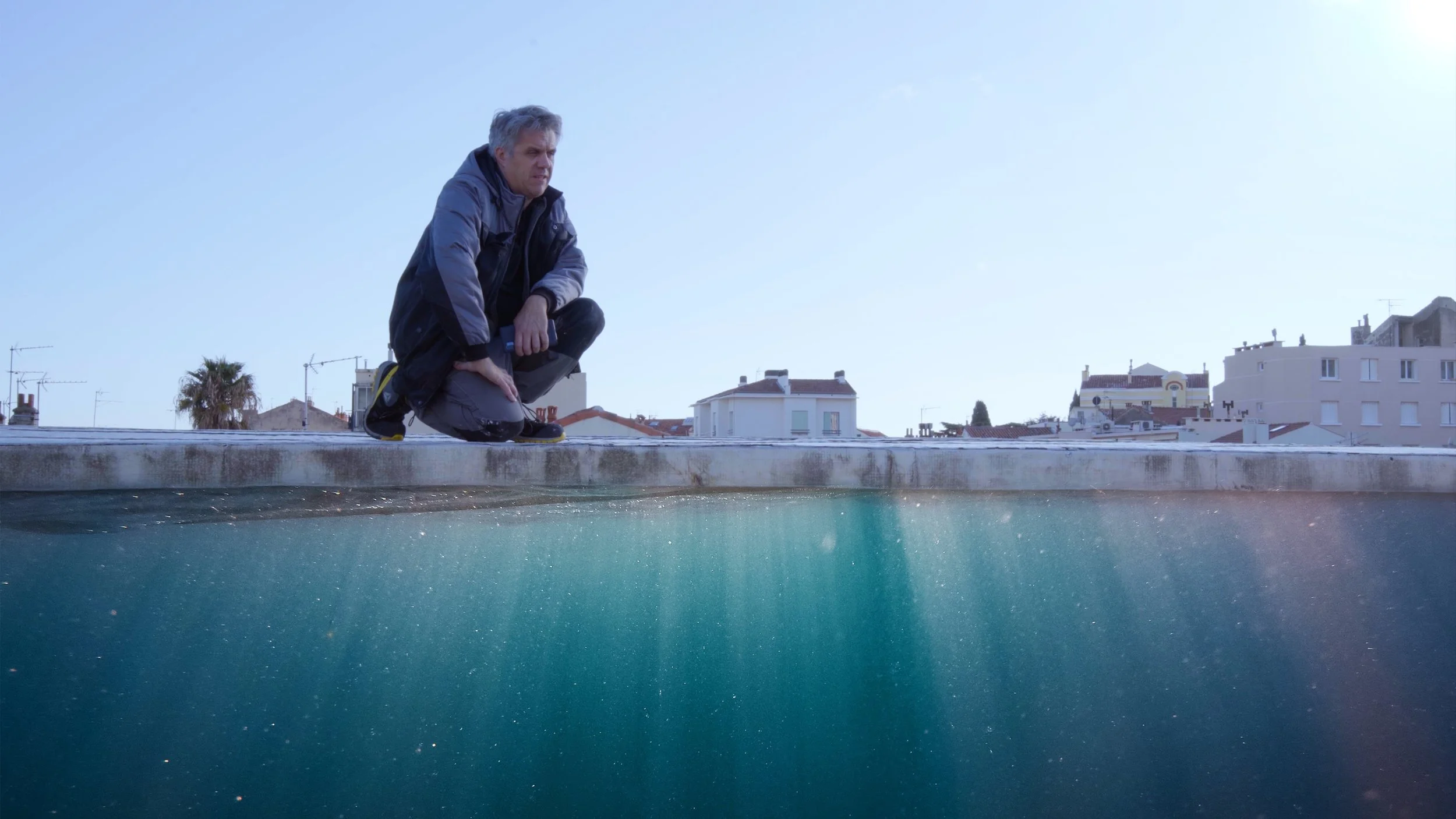 Un homme en veste sombre, accroupi sur un toit, regarde l'eau en dessous, avec des bâtiments résidentiels en arrière-plan.