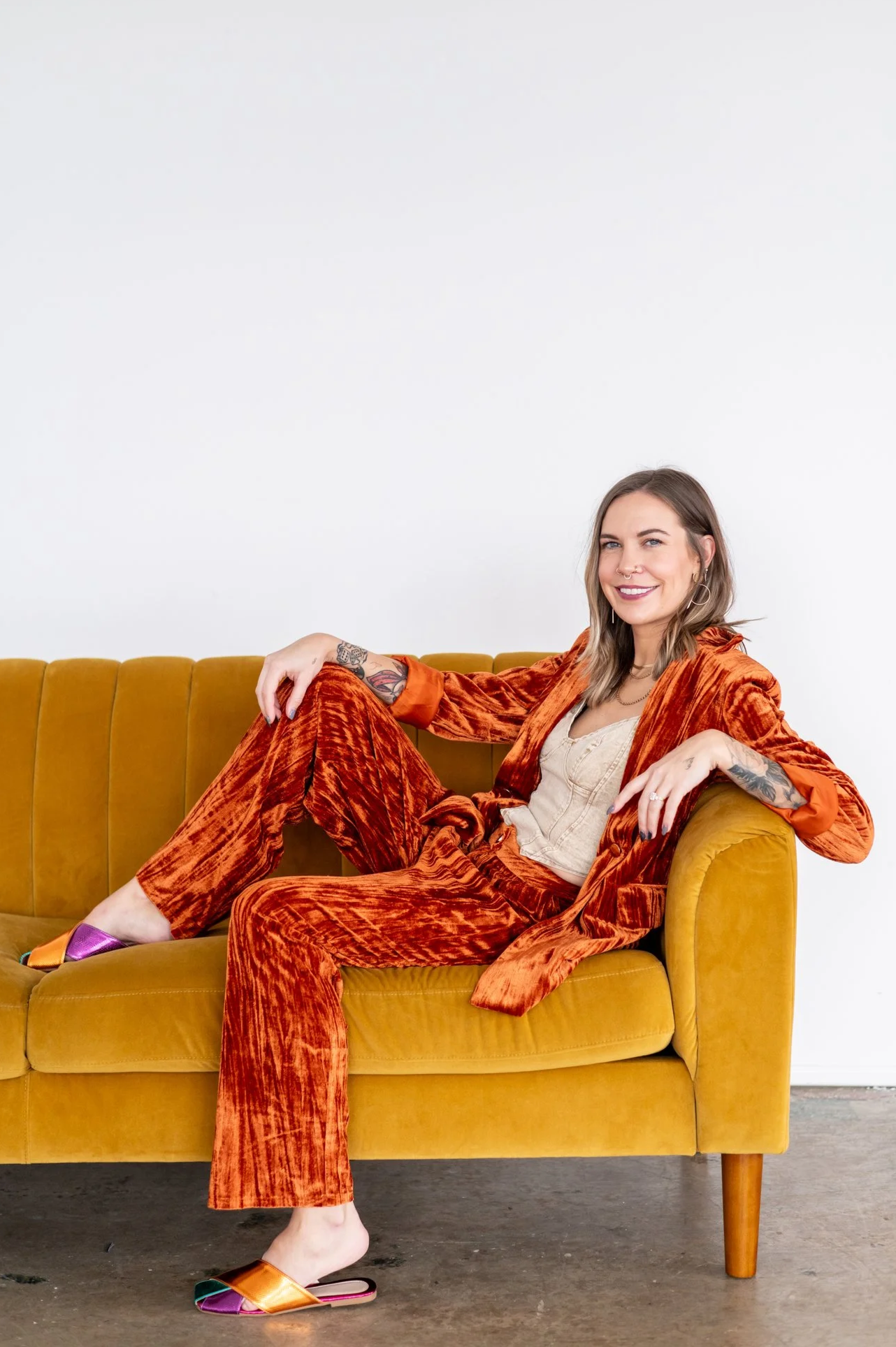 A woman with tattoos and shoulder-length hair sitting relaxed on a mustard yellow couch against a white wall, wearing an orange velvet suit, a beige top, and multicolored slippers.