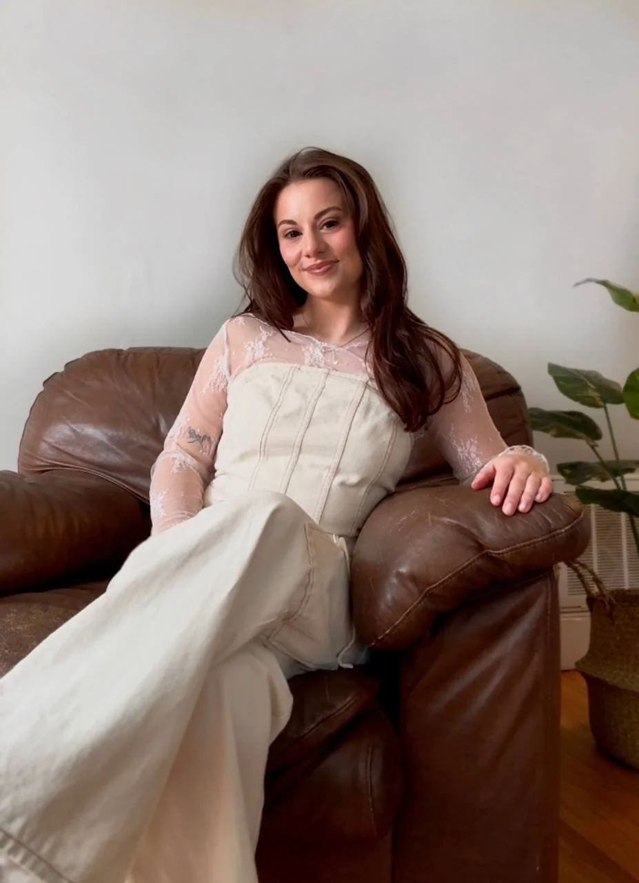 A young woman with long brown hair sitting on a brown leather couch, smiling at the camera, in a minimally decorated room with a white wall and a potted plant nearby.