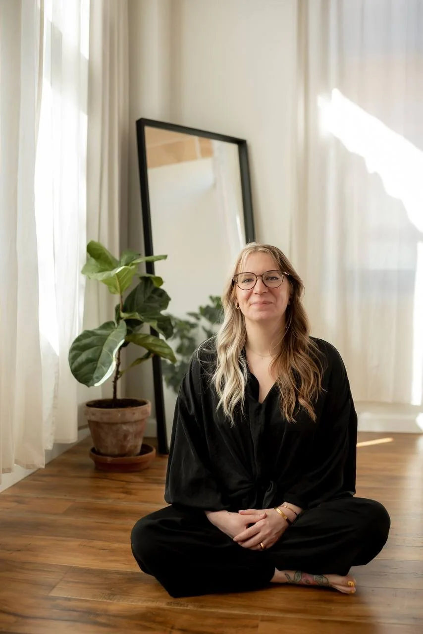 A woman with long blonde hair, glasses, and a black outfit sitting cross-legged on wooden floor in a bright room with white curtains. There is a potted plant and a mirror behind her.