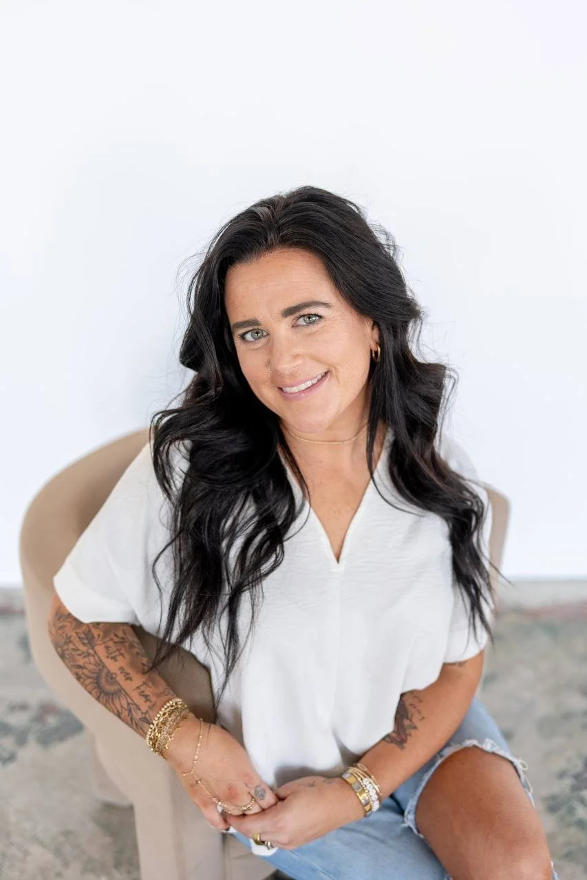 A woman with long black hair, wearing a white top and denim shorts, sitting on a beige chair against a plain white wall.