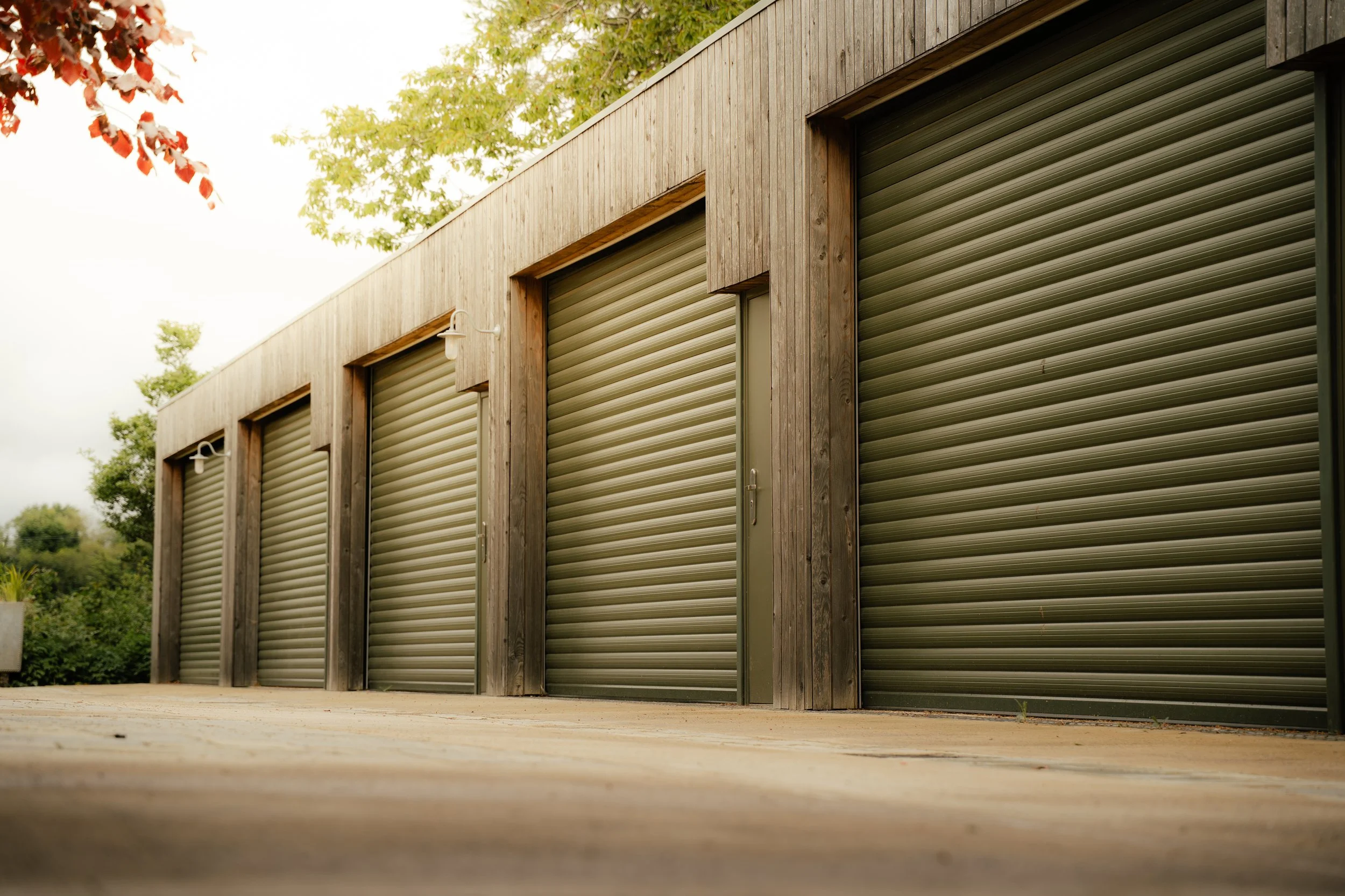 Row of commercial garage doors made of horizontal metal slats, framed by wooden panels, with outdoor lighting fixtures above some doors, on a concrete surface with trees in the background.