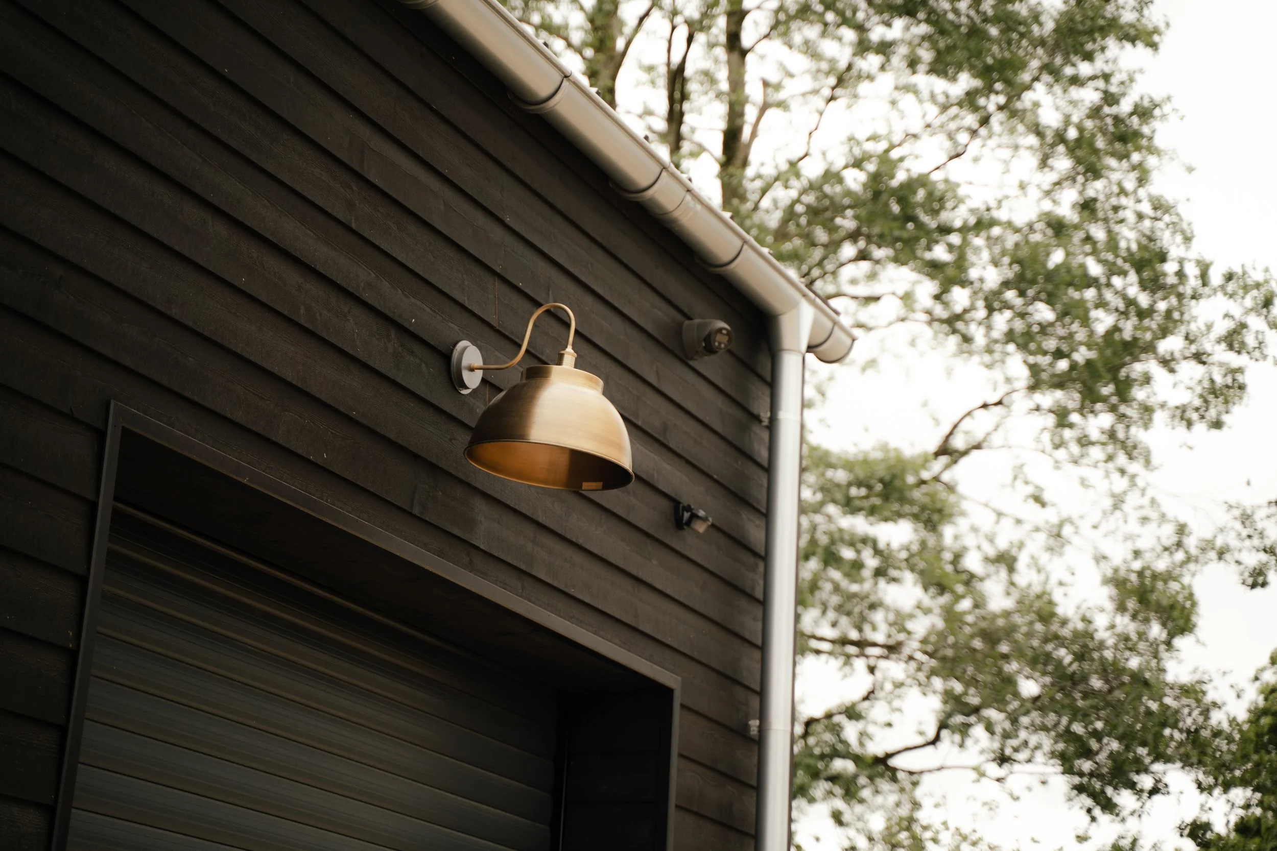 Exterior view of a black wooden house with a gold outdoor wall lamp, a rain gutter, security camera, and a background of leafy trees.
