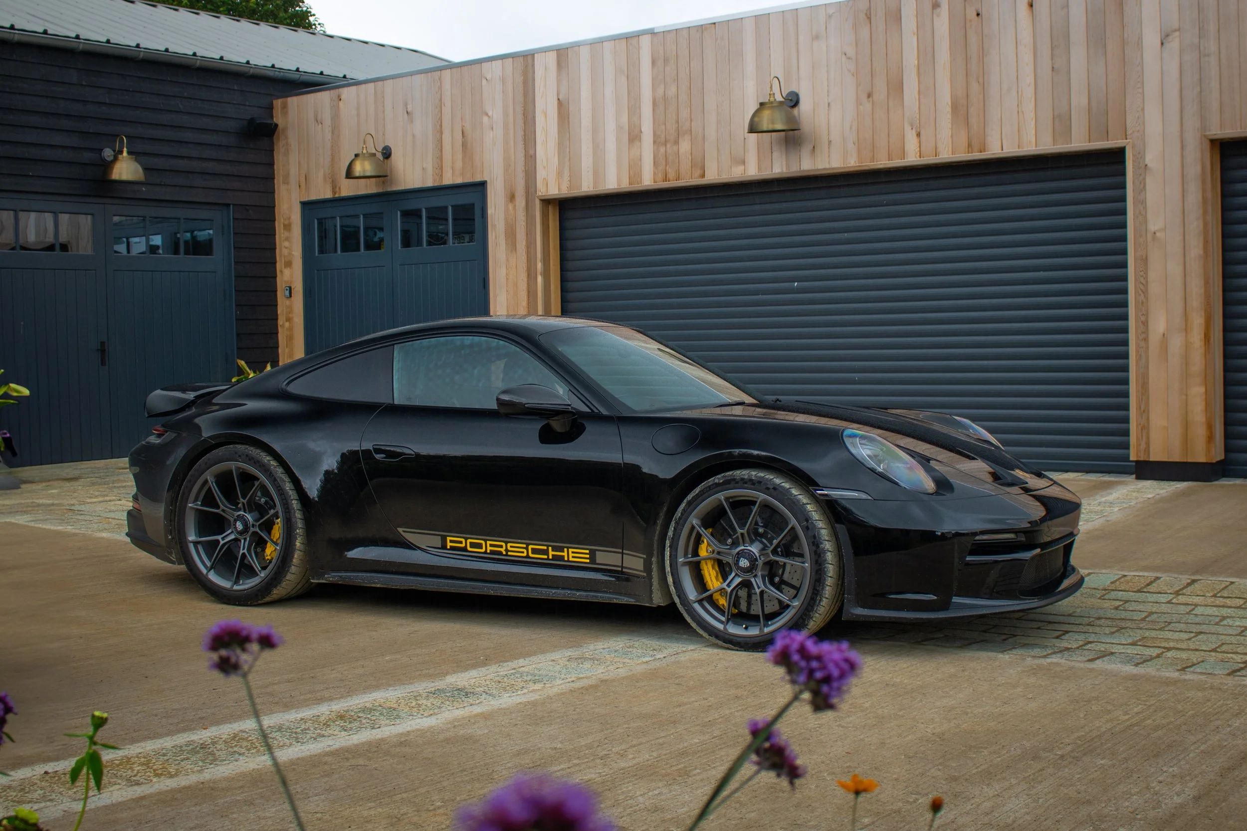 A black Porsche sports car parked in front of a modern building with wood-paneled walls and two closed garage doors. Flower plants with purple flowers are in the foreground.