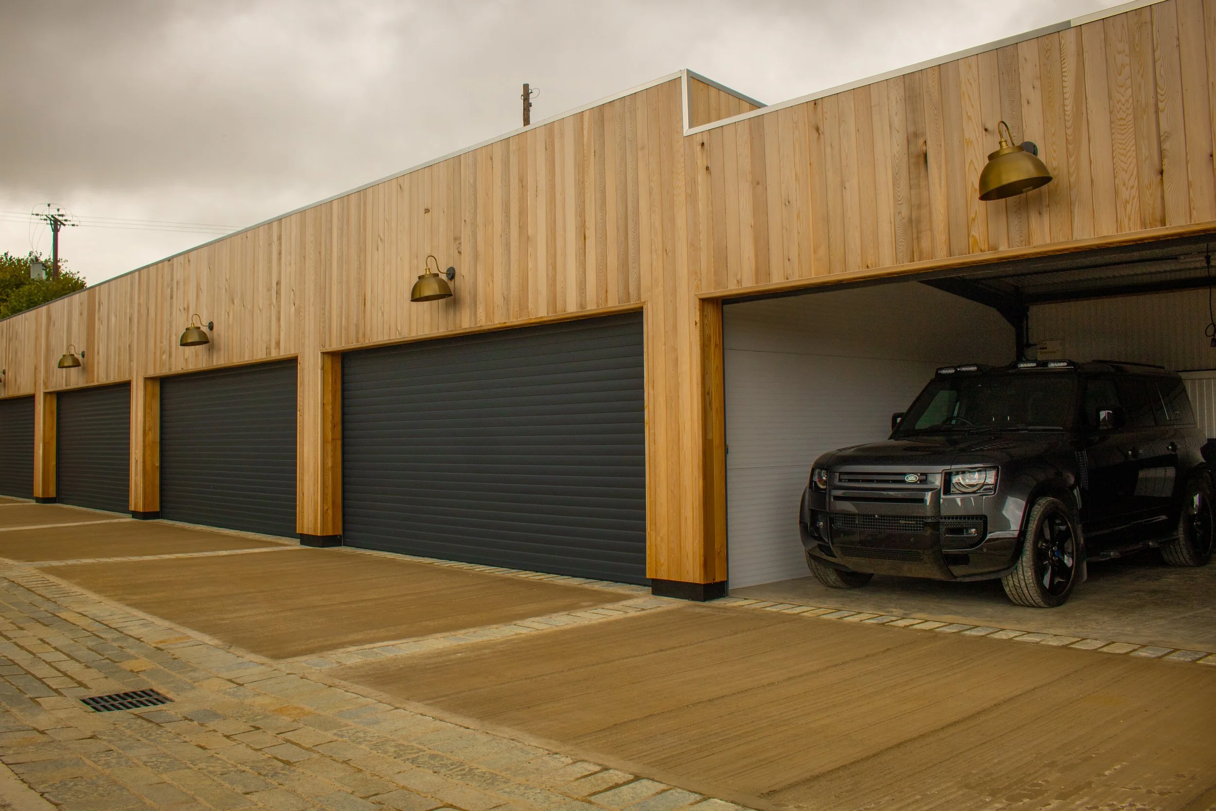A row of storage units with black garage doors and wooden siding, one of which has a black SUV parked inside, and a concrete driveway in front.