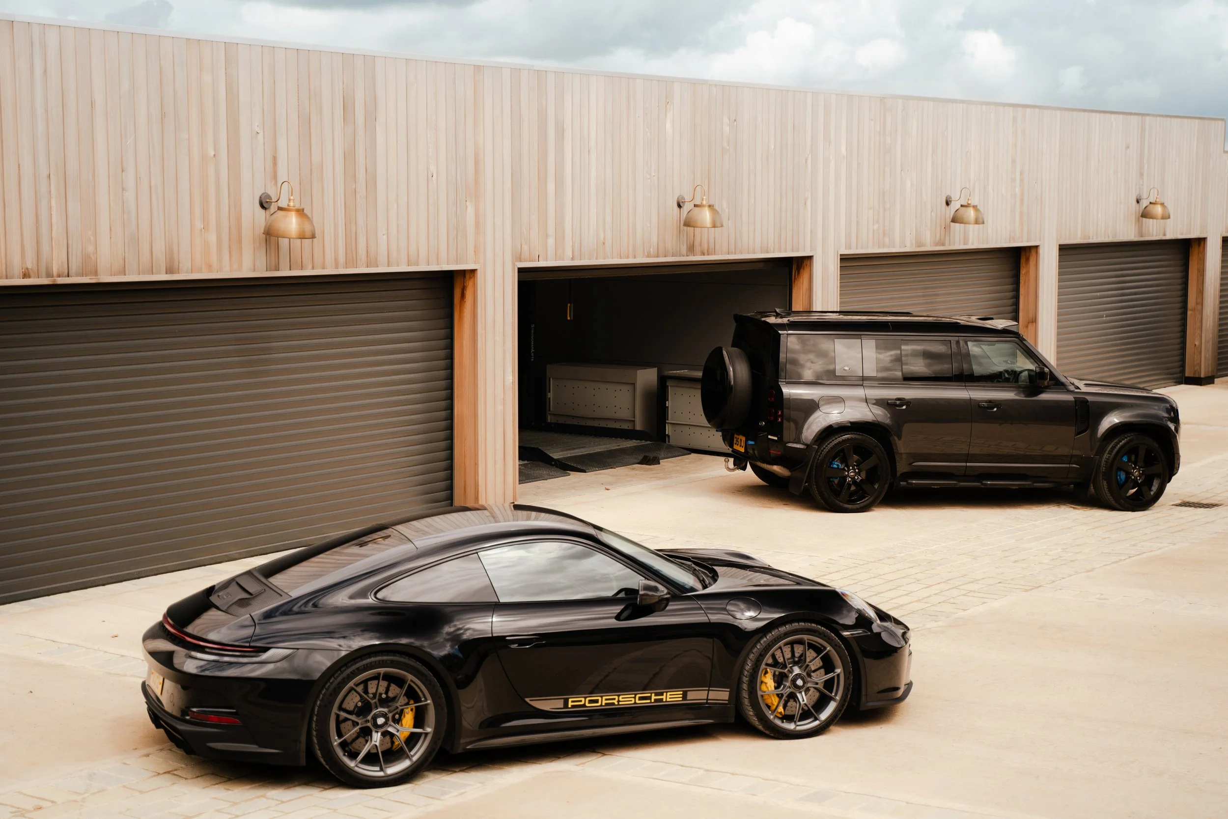 Black Porsche sports car parked in front of a wooden building with garage doors, with another vehicle partially visible at the bottom of the image.