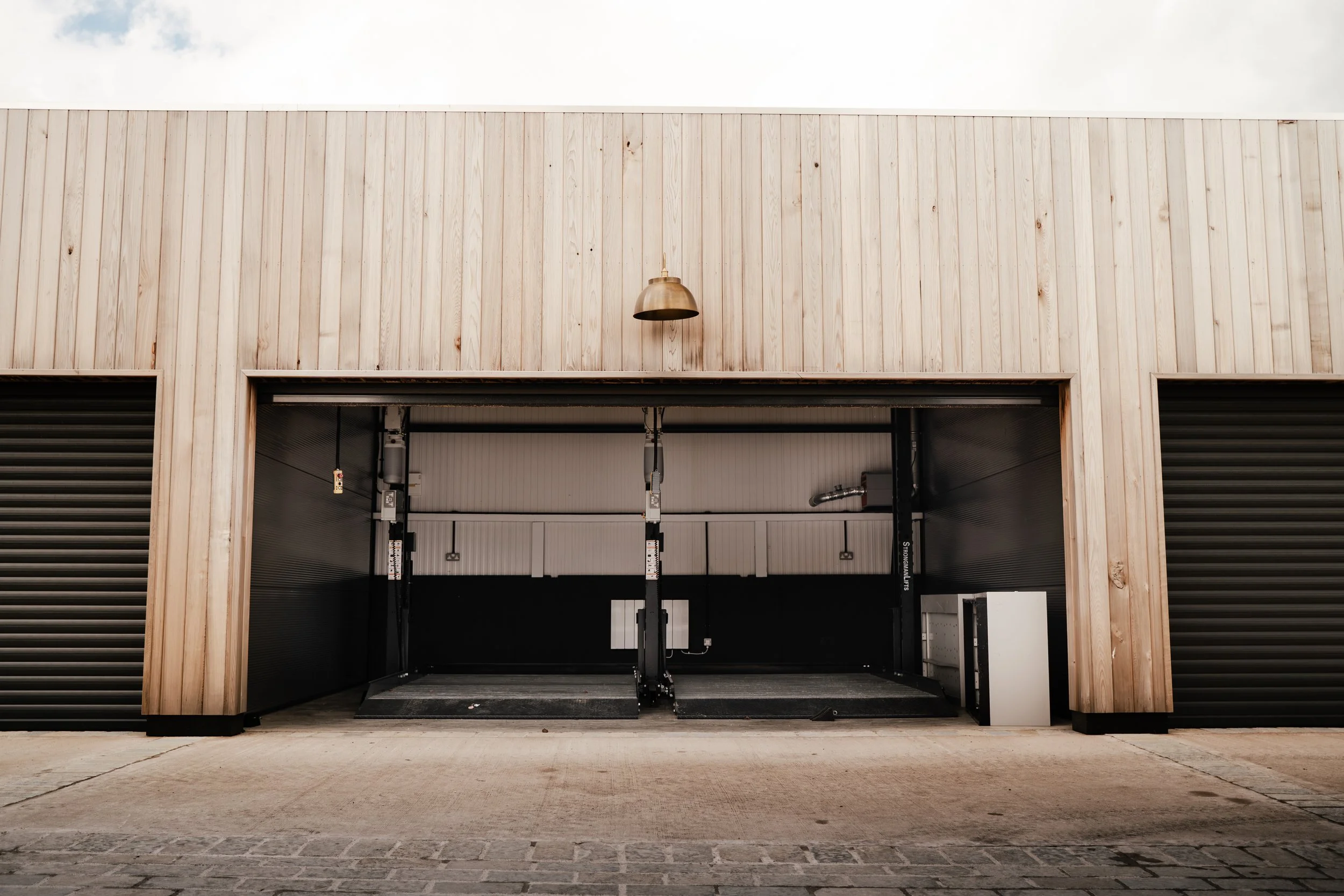 Open garage with black lifts inside, beige wooden exterior, and a single bronze-colored exterior light fixture.