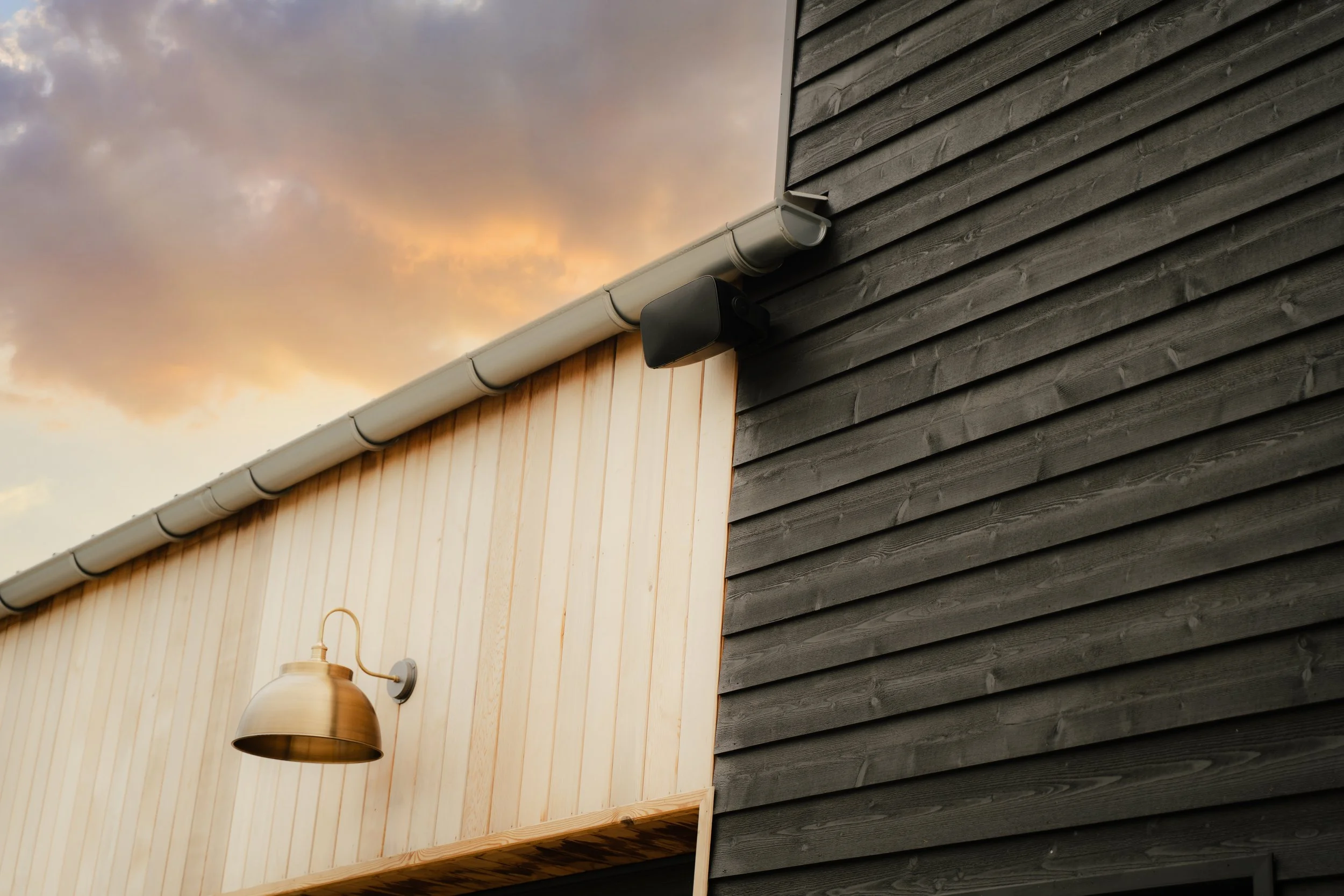 Exterior view of a building with dark gray wooden siding on one side and light-colored wood siding on the other, featuring a black outdoor security camera and a wall-mounted brass outdoor light fixture, under a cloudy evening sky.