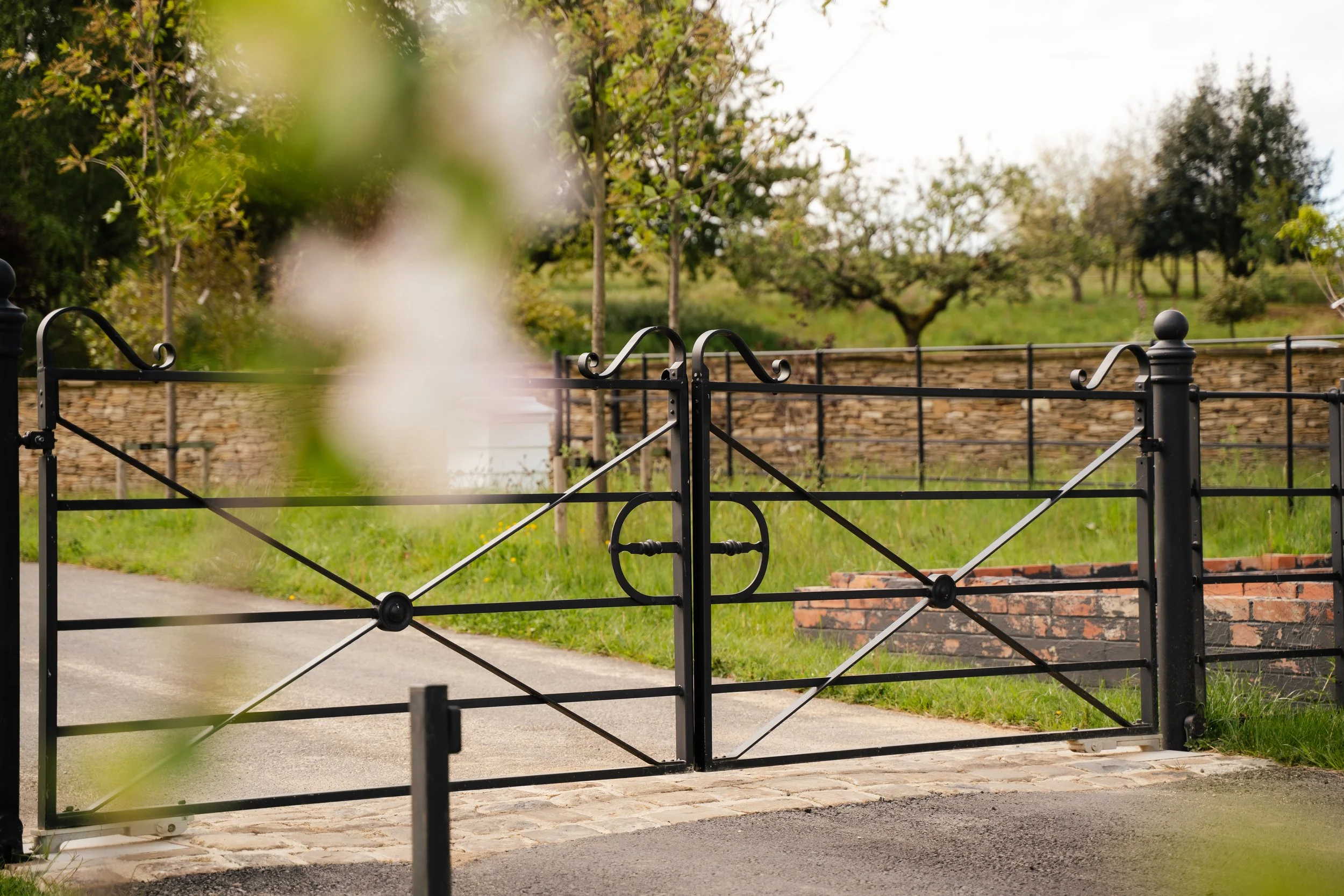 Black metal gate with decorative swirls, partially open, in front of a stone pathway leading into a yard with green grass and trees, with a brick wall background and overcast sky.