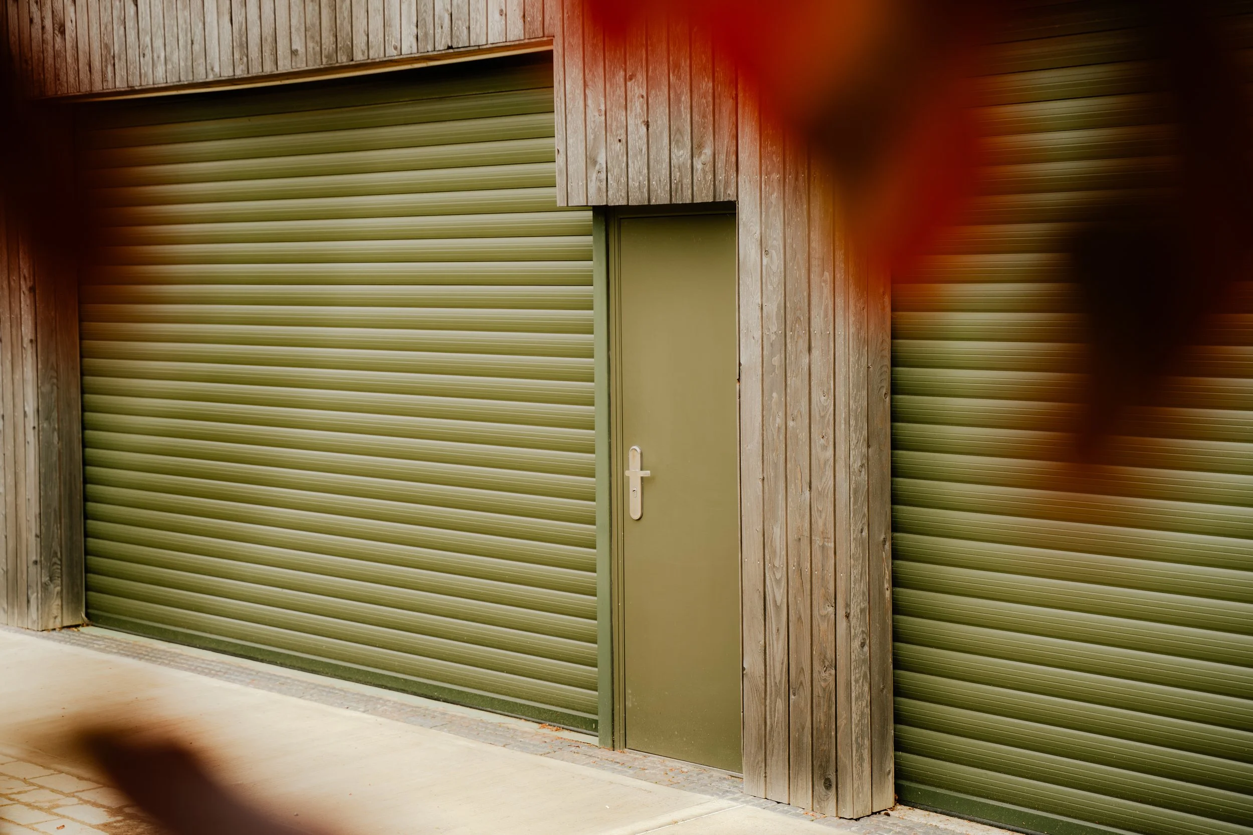 Green rolling shutter door set in wooden wall with a small door to the right, located outdoors on a concrete ground.