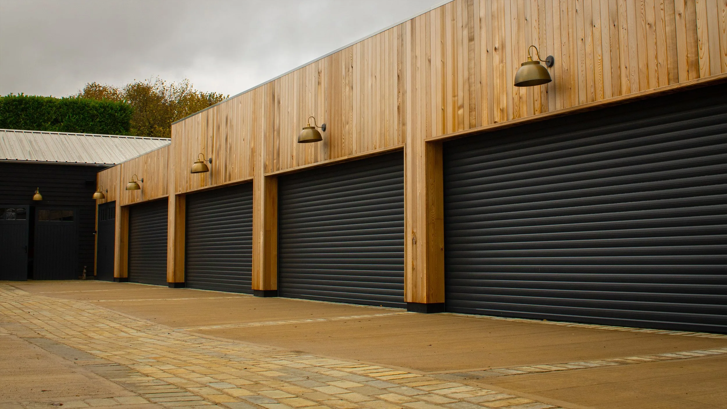 Row of black garage doors with wooden siding and outdoor light fixtures above each door, in a commercial or industrial area.