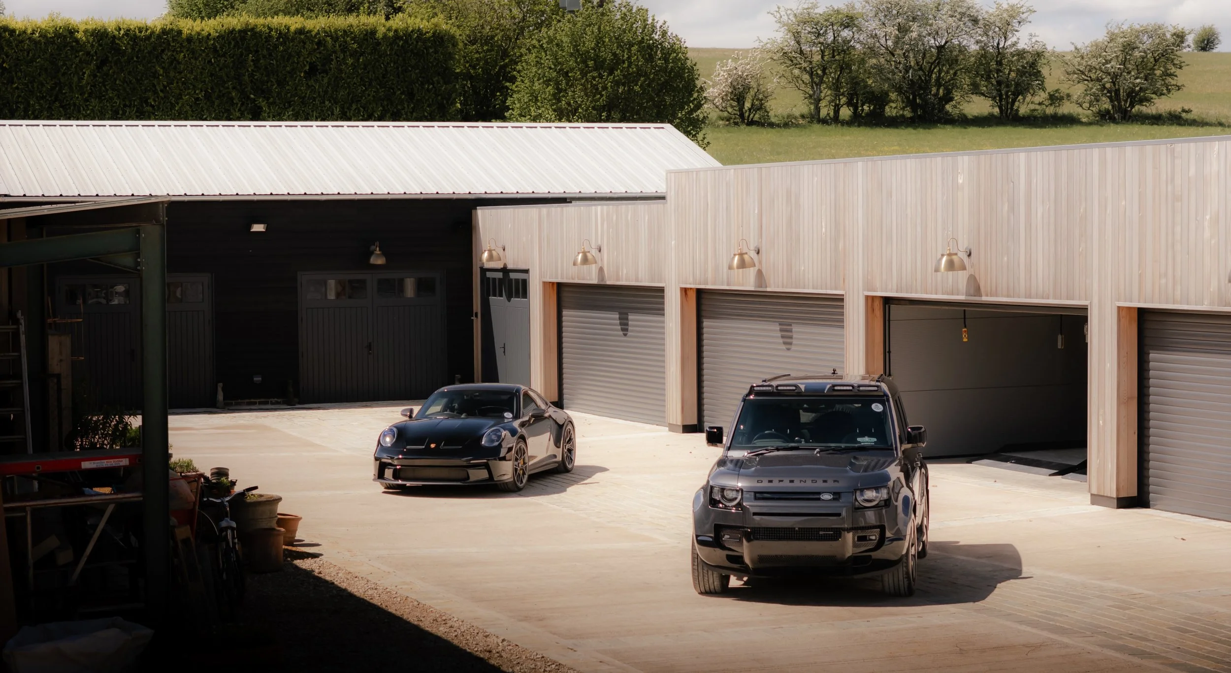 A garage with three roll-up doors, two of which are open. Two cars are parked outside: a black sports car and a gray utility vehicle. There is open space around the garage with trees and greenery in the background.