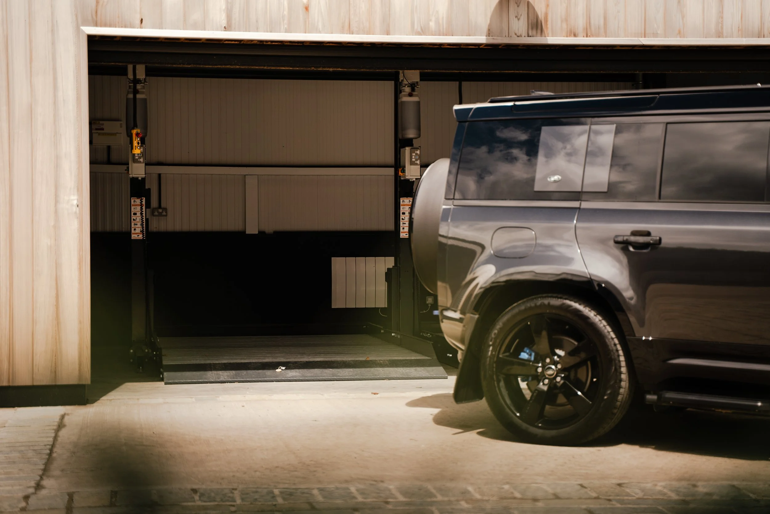 The image shows a garage with an electric vehicle parked just outside it, and the garage door is open, revealing the interior with mechanical lift equipment.