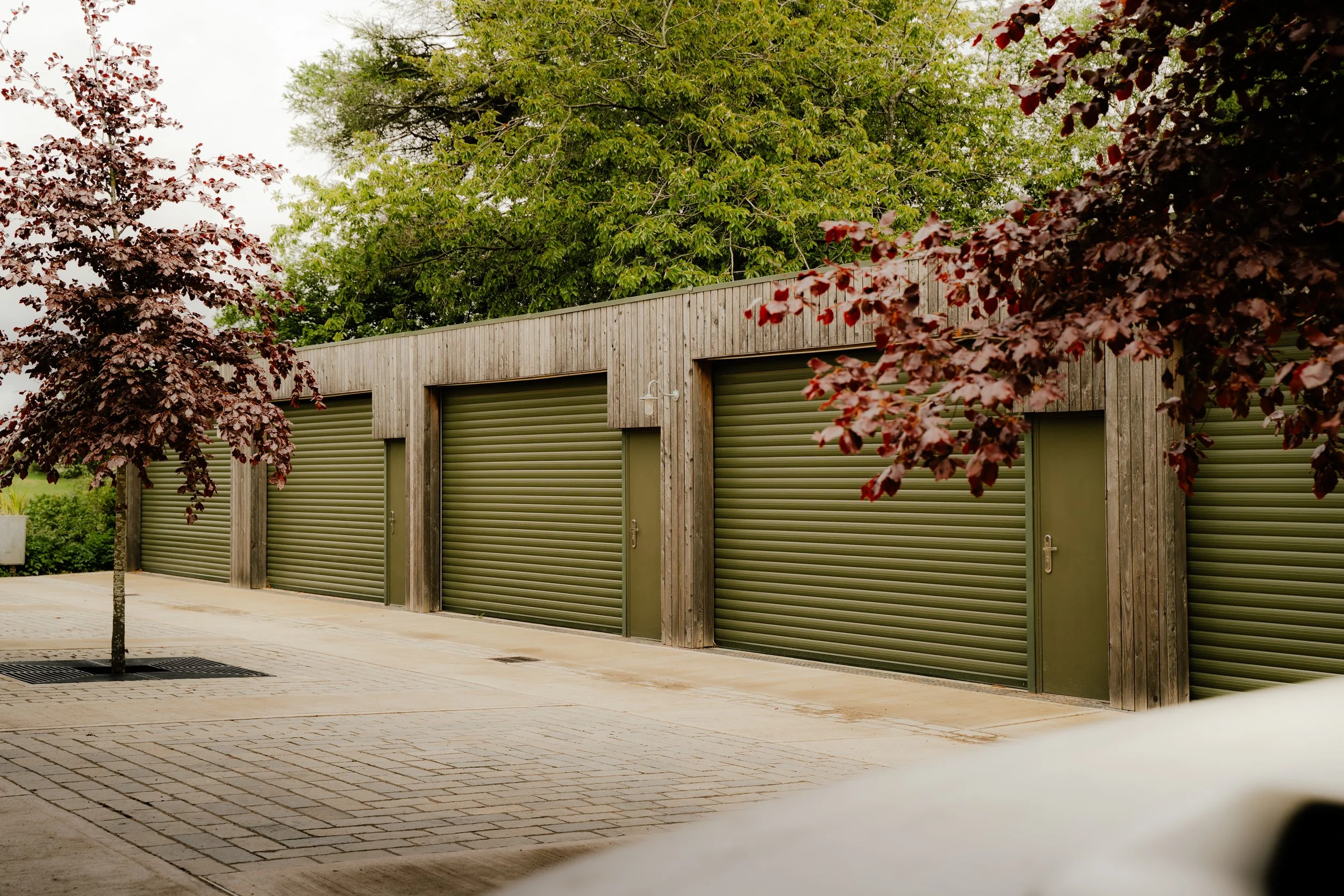 A row of green rolling garage doors in a modern wooden building, with trees in the background and a paved sidewalk in front.