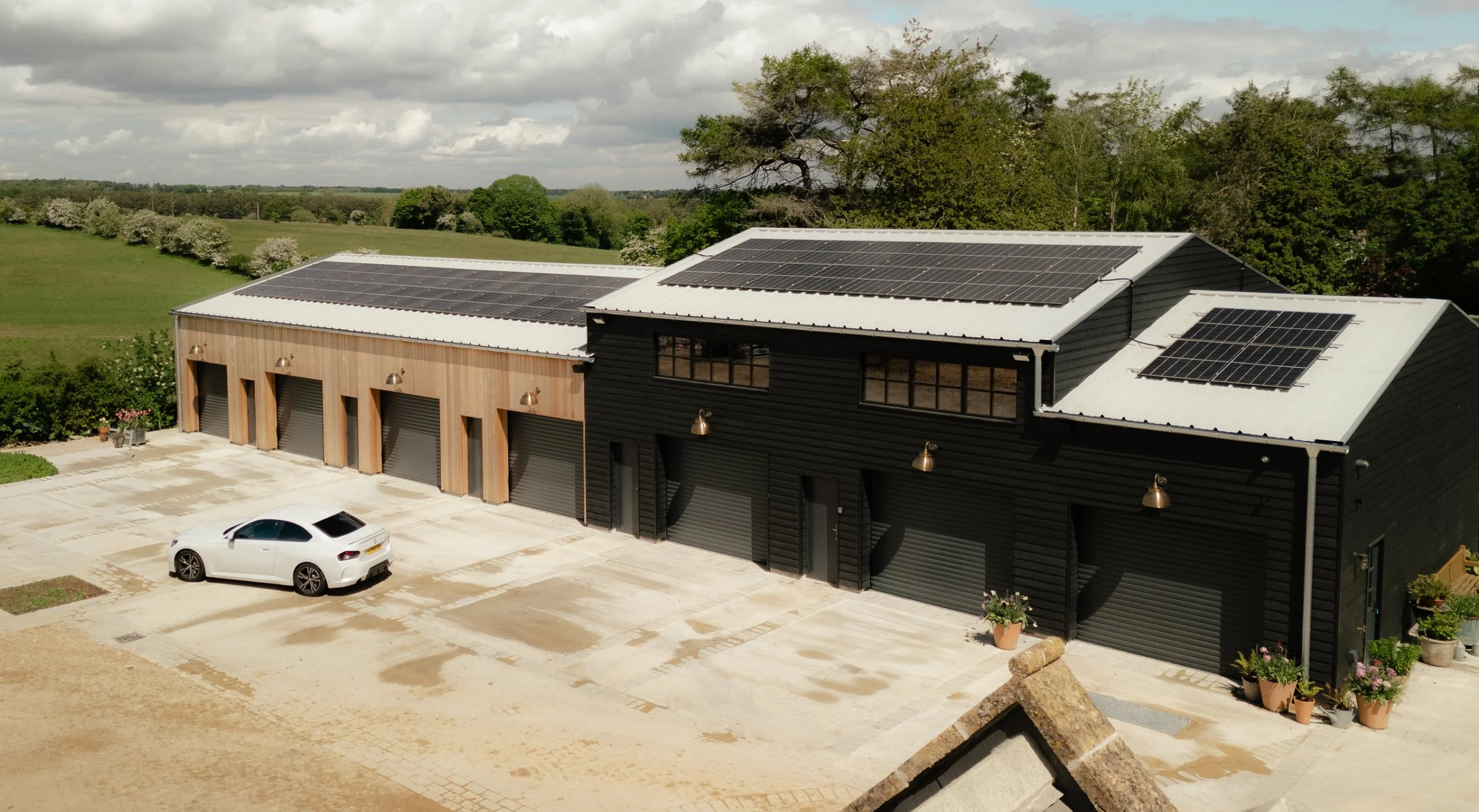 A building with solar panels on the roof, black and wood-paneled exterior walls, a parking lot with a white car, and surrounding greenery including trees and grass, under a partly cloudy sky.